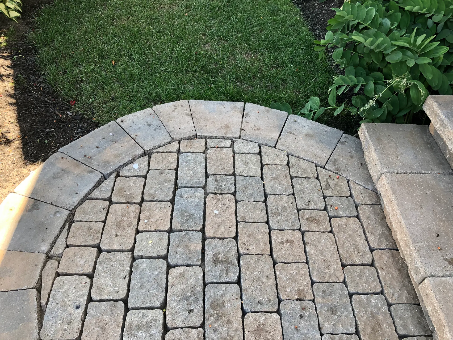 Outdoor landing with grey interlocking pavers, a rounded border, stone steps, grass, and surrounding landscaping.