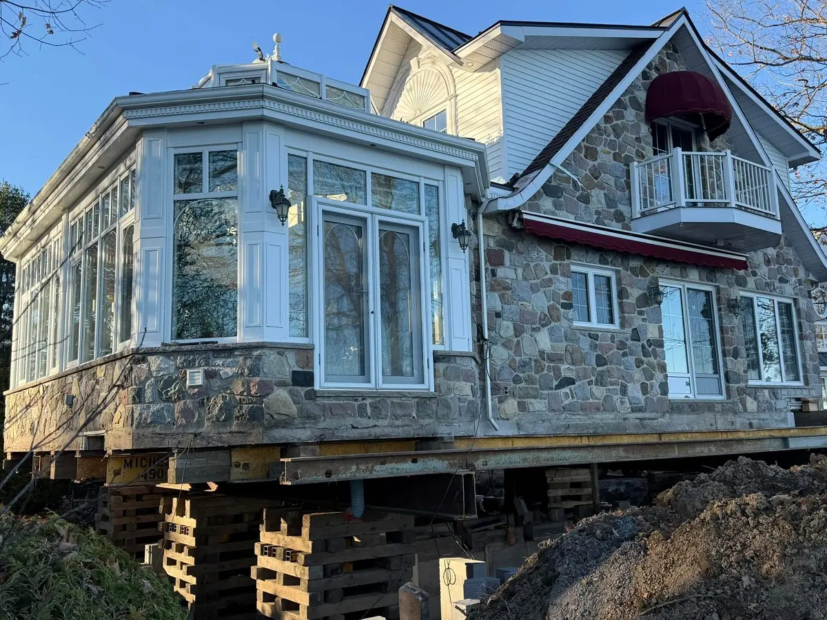 Raised stone house under construction with glass sunroom, white balcony, red awnings, patio doors and exposed foundation