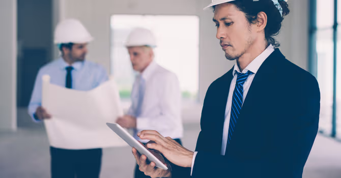 Professionnel de la construction avec casque utilisant une tablette pendant que des collègues consultent des plans sur un chantier intérieur.