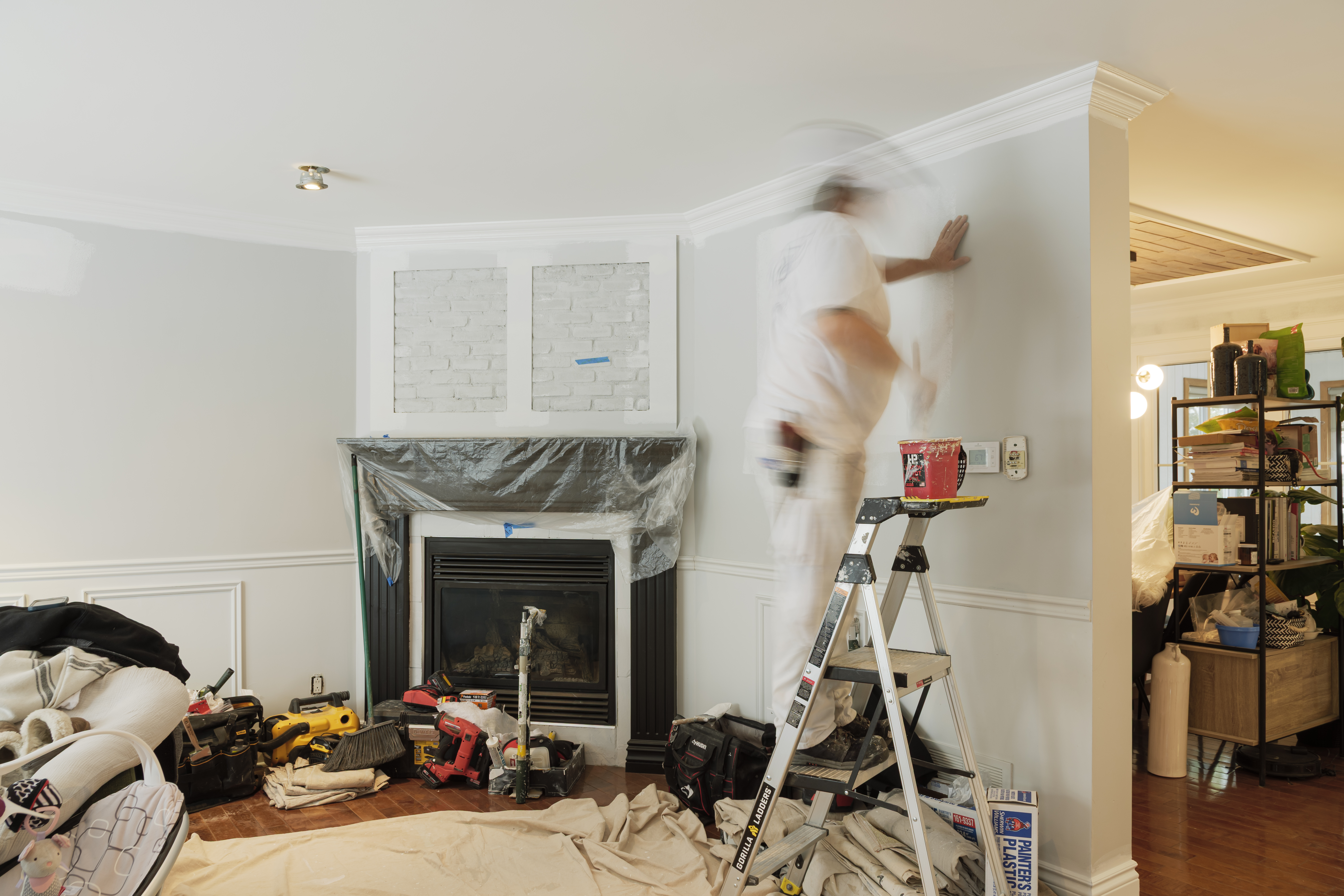 Travailleur peinturant les murs d’un salon en rénovation, avec moulures décoratives, foyer protégé, escabeau, outils et bâches de protection.