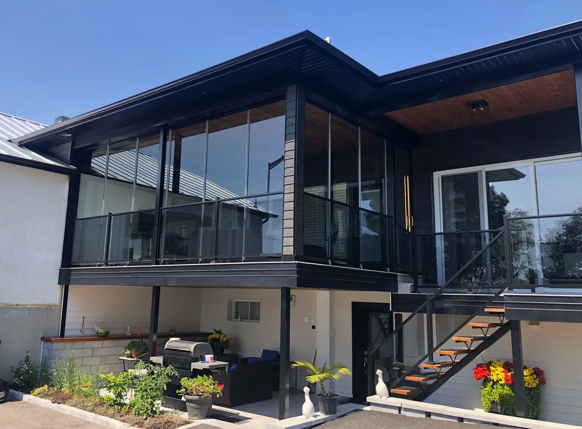 Maison moderne avec balcon vitré, structure noire, escalier extérieur et espace terrasse aménagé au rez-de-chaussée