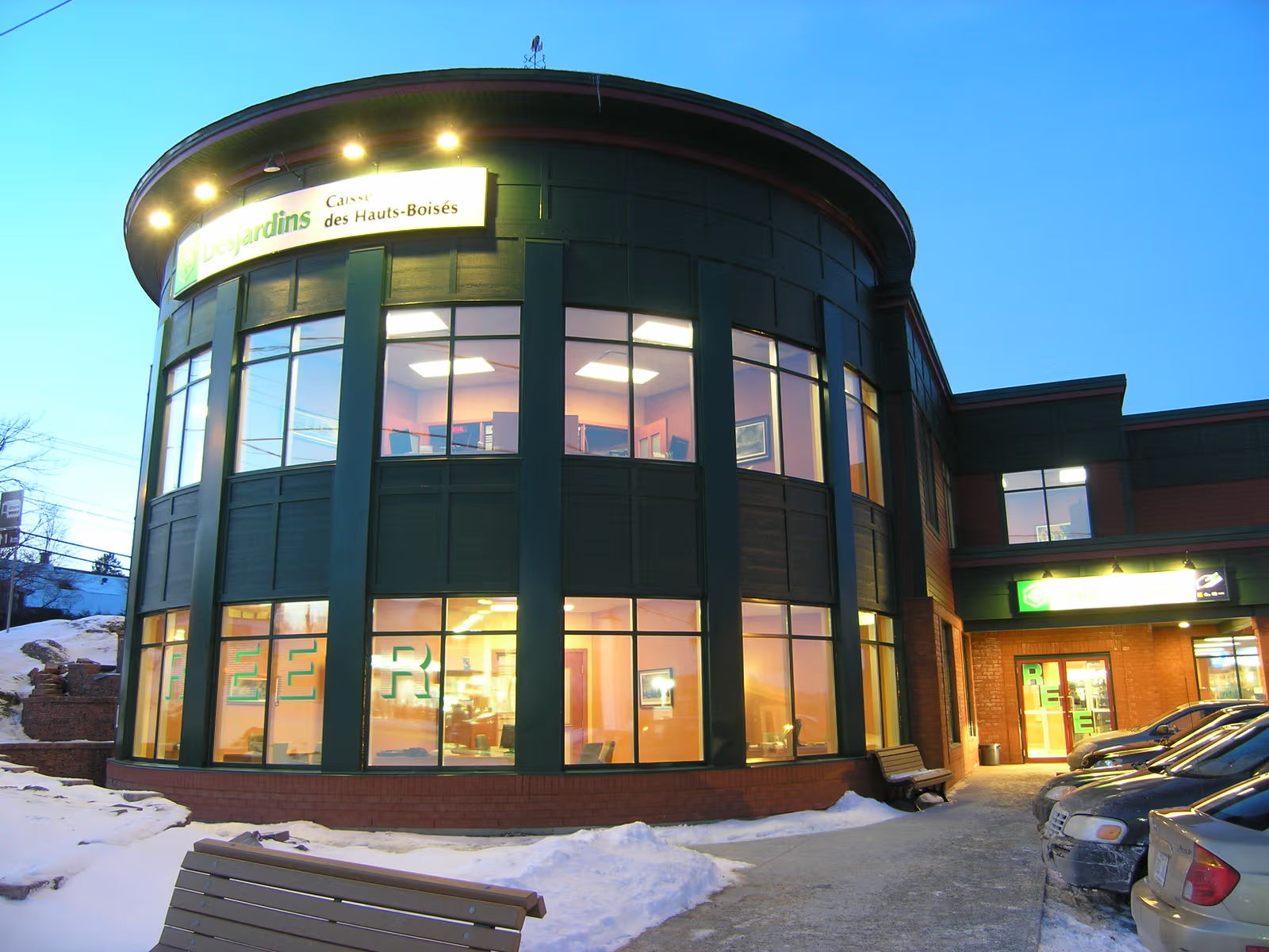 Bâtiment commercial moderne éclairé en soirée, avec grandes fenêtres vitrées, entrée principale, stationnement et neige autour de la façade.