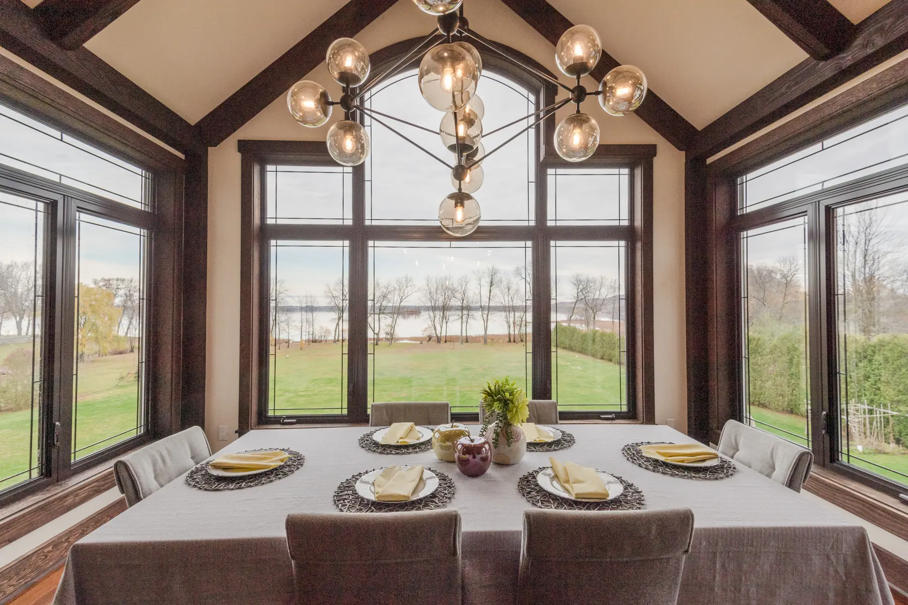 Salle à manger élégante avec une grande table recouverte d'une nappe beige, un lustre design, des fenêtres panoramiques offrant une vue sur un grand jardin et un lac en arrière-plan.