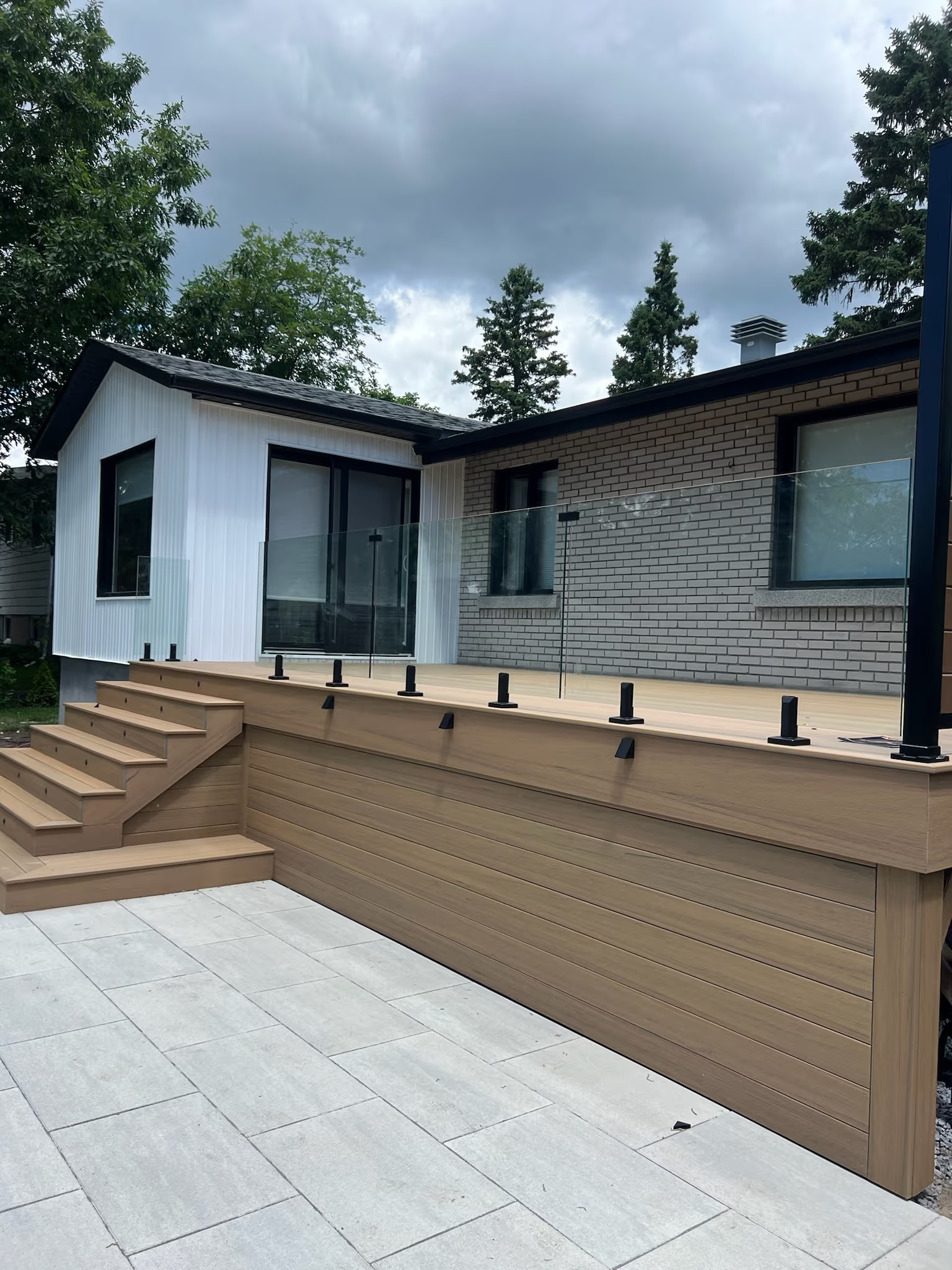 Terrasse surélevée en bois composite avec escalier, garde-corps en verre et patio en dalles devant une maison contemporaine en brique et revêtement blanc