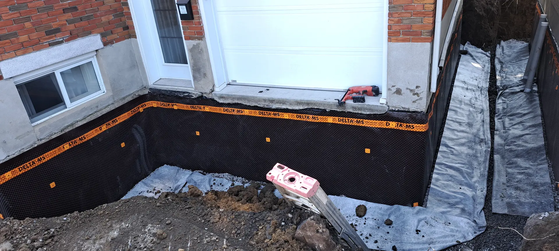 Fondation de maison excavée avec membrane d’imperméabilisation noire et drain installé près d’une porte de garage