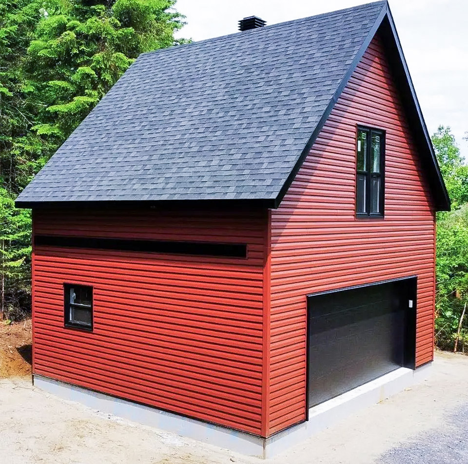 Garage détaché à deux étages avec revêtement rouge, porte noire, fenêtres assorties et toiture à forte pente en bardeaux foncés.