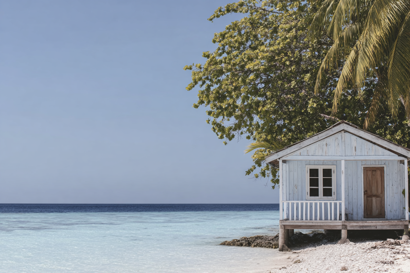 Small white wooden cabin on stilts beside a tropical beach, with turquoise sea, clear sky, palm trees and lush greenery