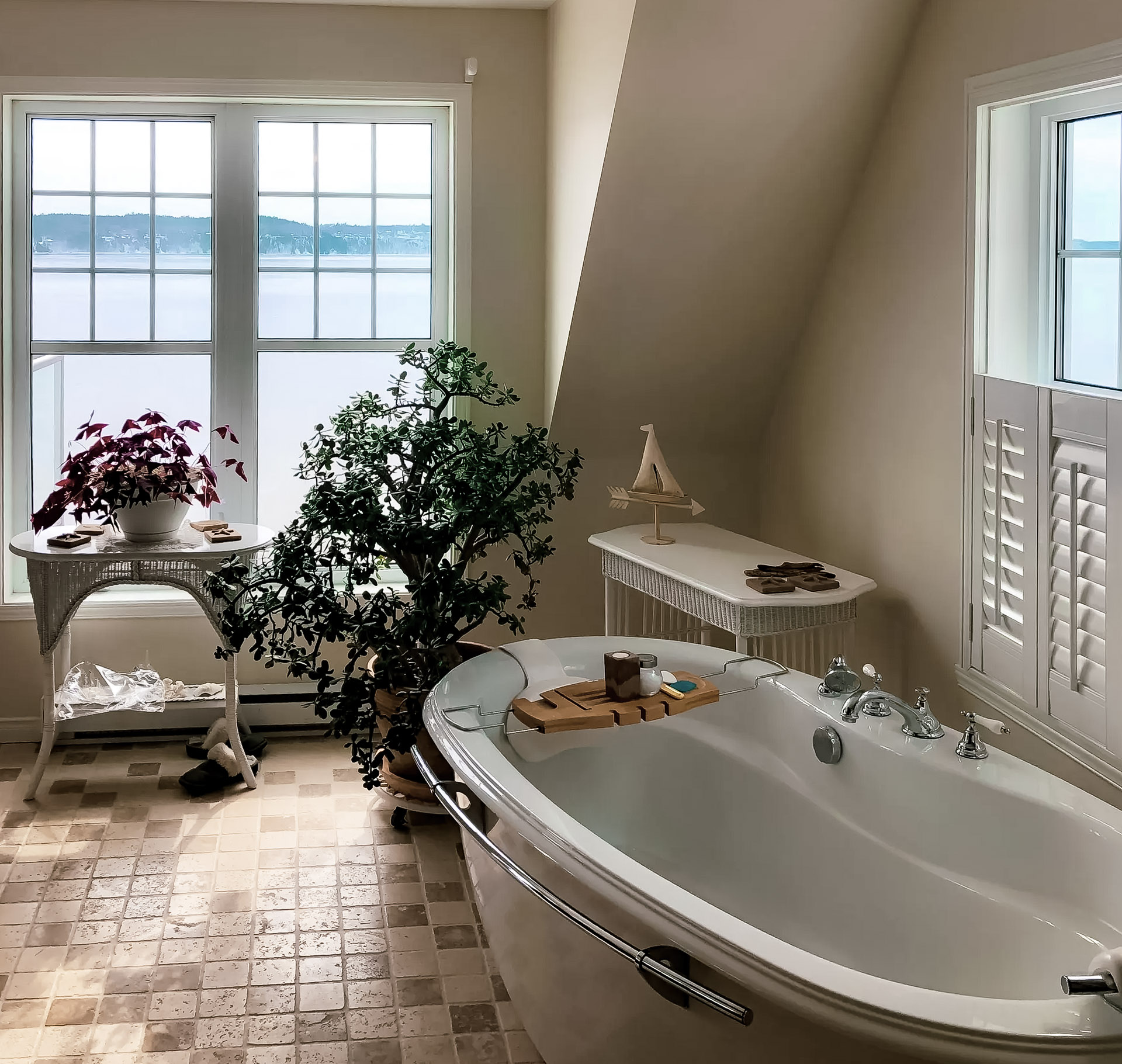 Bright bathroom with a freestanding bathtub, chrome fixtures, indoor plants, wicker furniture, and large windows overlooking a waterfront landscape.