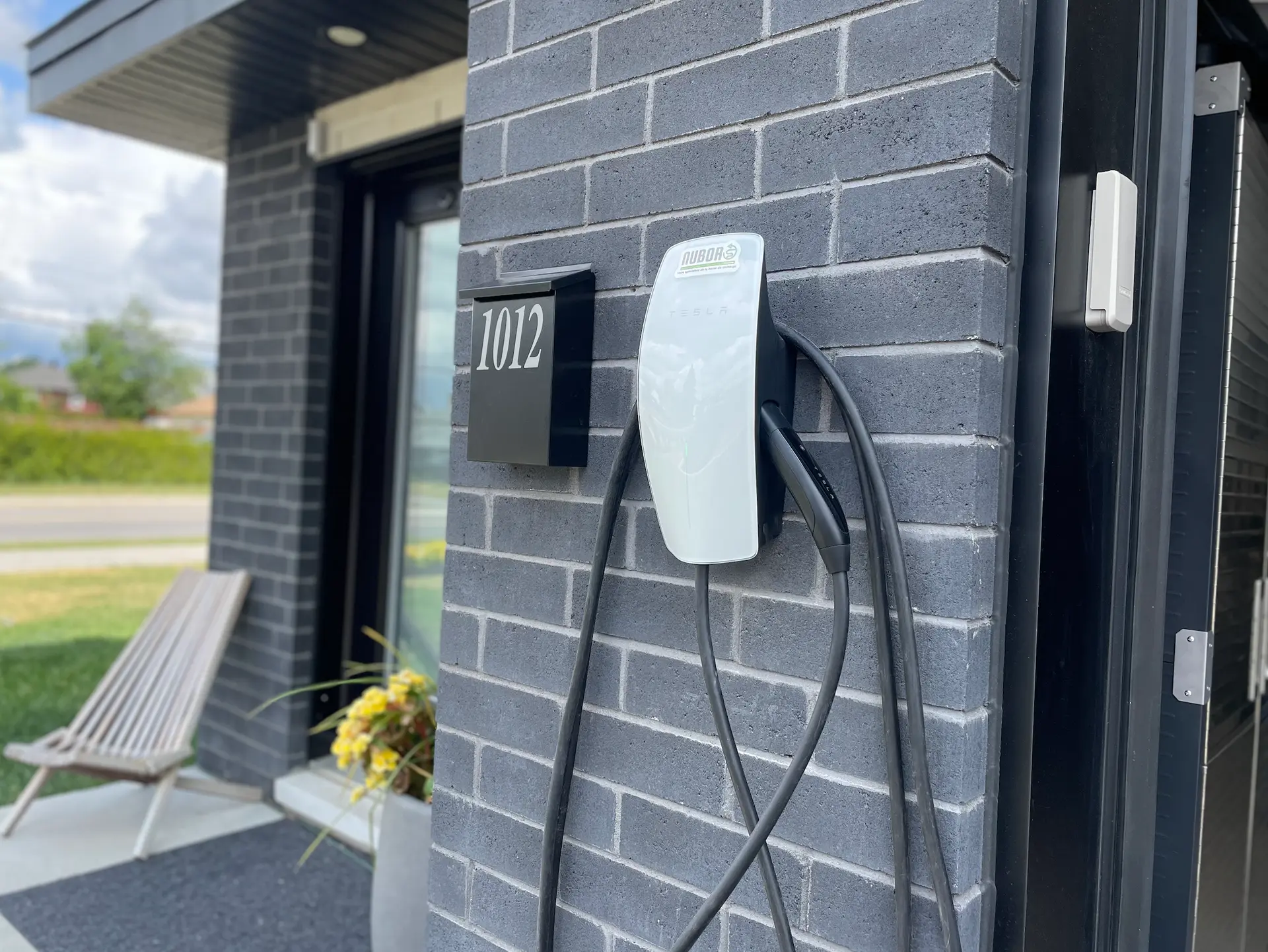 Wall-mounted electric vehicle charging station installed on a gray brick exterior with charging cable, modern entrance, and house number 1012
