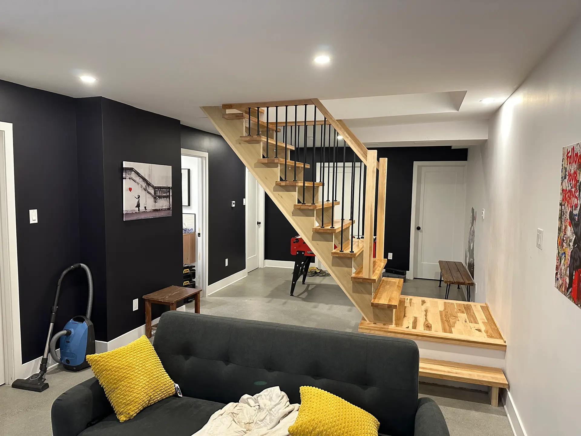 Renovated basement with a wooden staircase, black and white walls, recessed lighting, grey sofa, and yellow cushions.