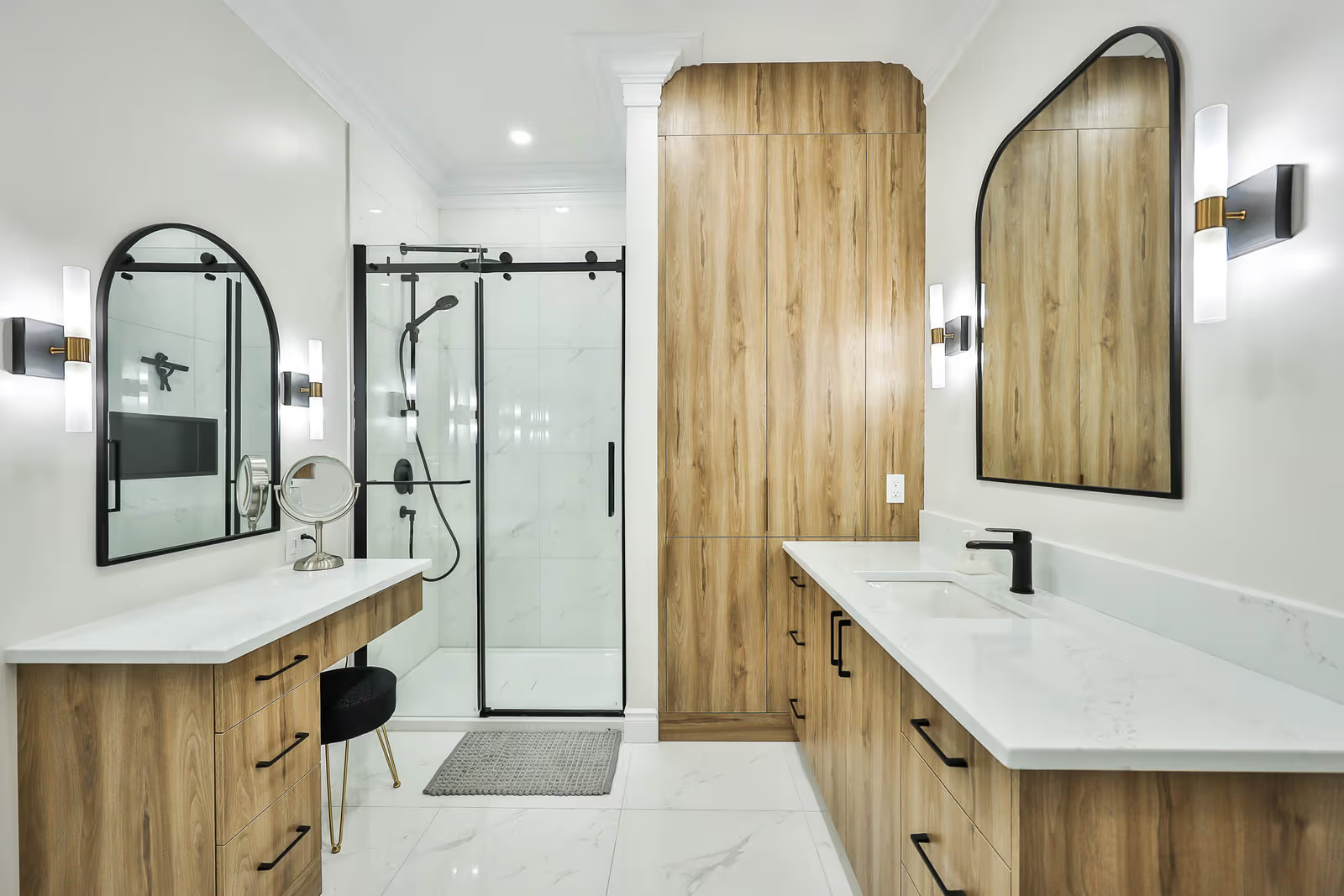 Large modern bathroom with light wood vanities, white countertops, arched mirrors, glass shower, black fixtures and wall lighting