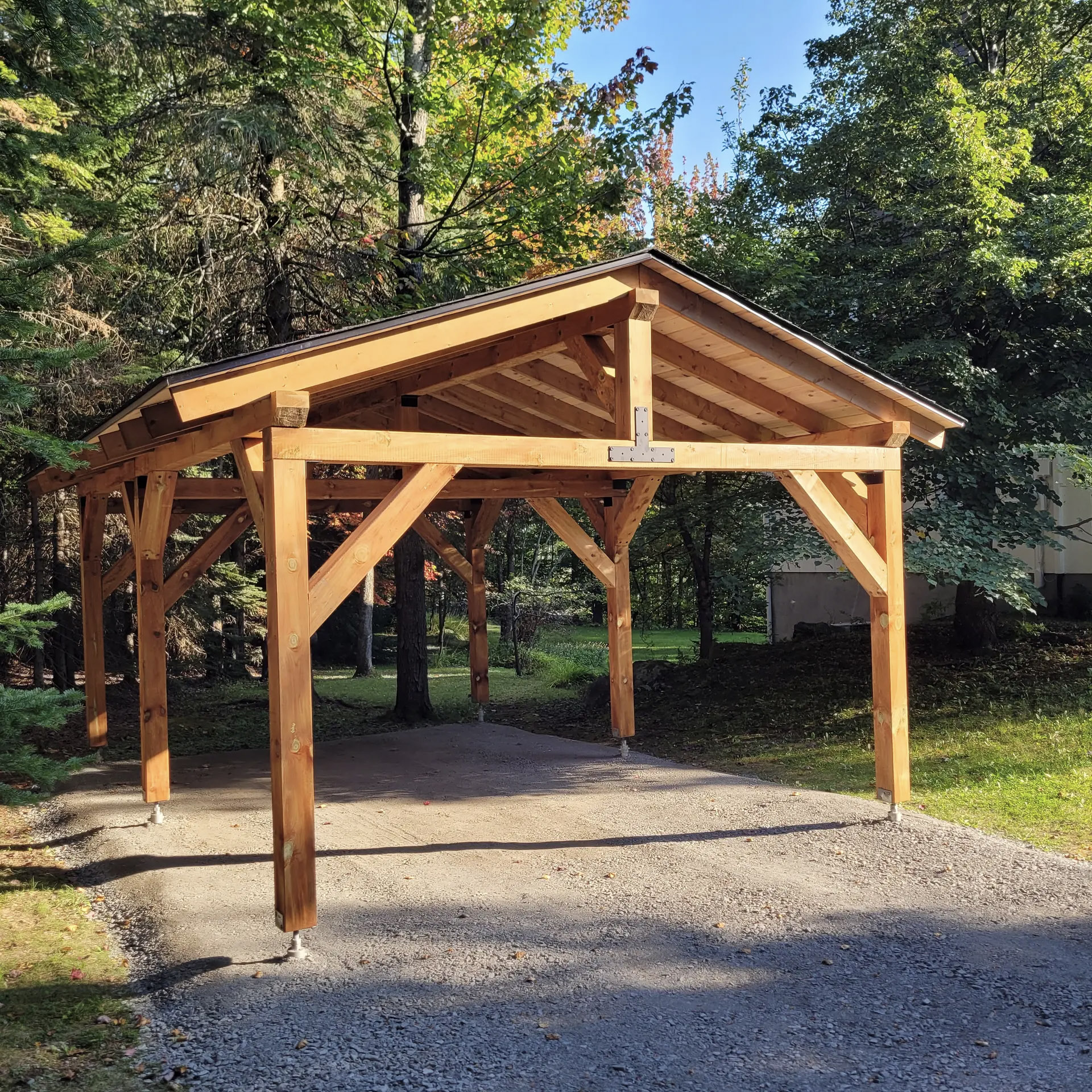 Abri extérieur en bois avec structure apparente, toiture inclinée et poteaux fixés sur une entrée en gravier entourée d’arbres.