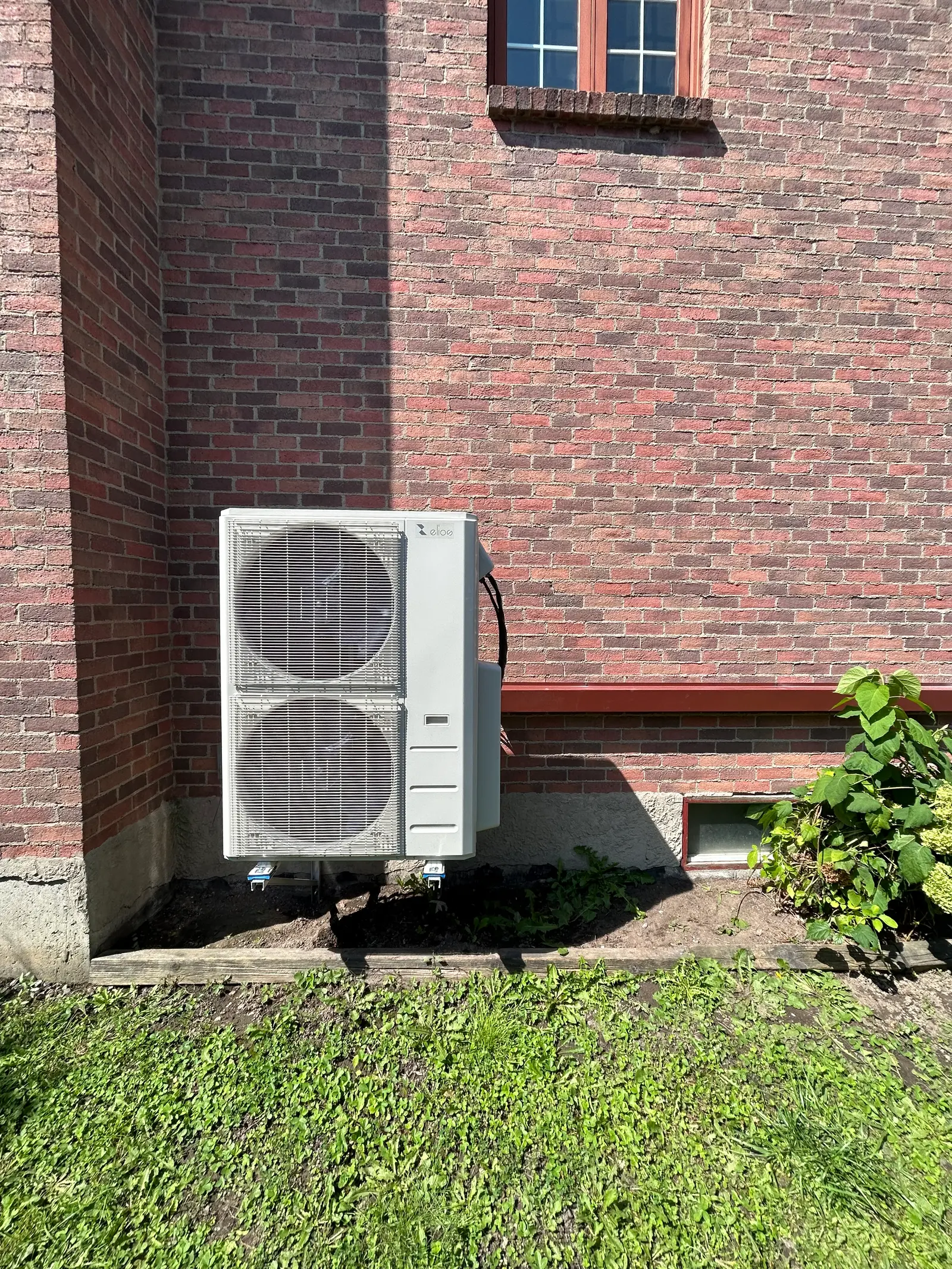Outdoor heat pump unit installed against a residential brick wall with ground vegetation