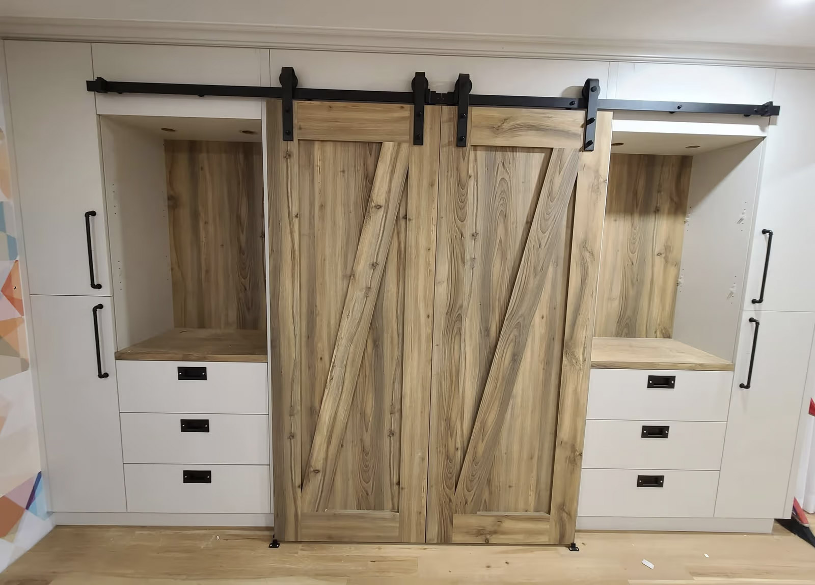 Portes coulissantes en bois clair sur rail noir, intégrées à un rangement blanc avec tiroirs et niches ouvertes au style rustique moderne.