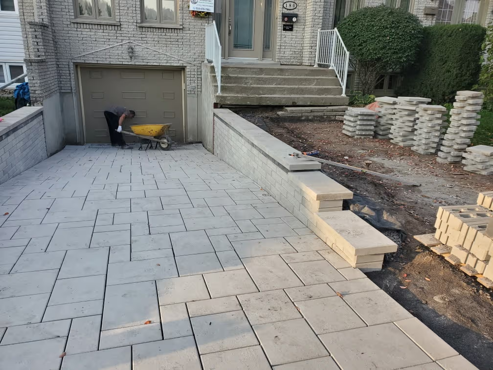 Grey interlocking paver installation in front of a house, with a garage entrance, retaining wall, stacked pavers, and a worker on site.