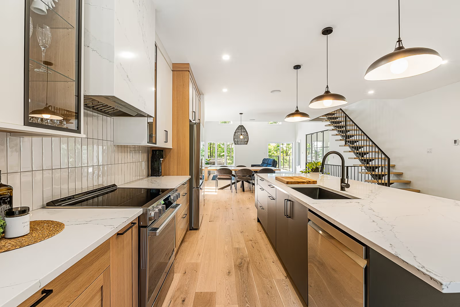 Cuisine moderne ouverte avec armoires en bois, îlot central foncé, comptoirs blancs veinés, luminaires suspendus, dosseret en céramique et escalier intérieur