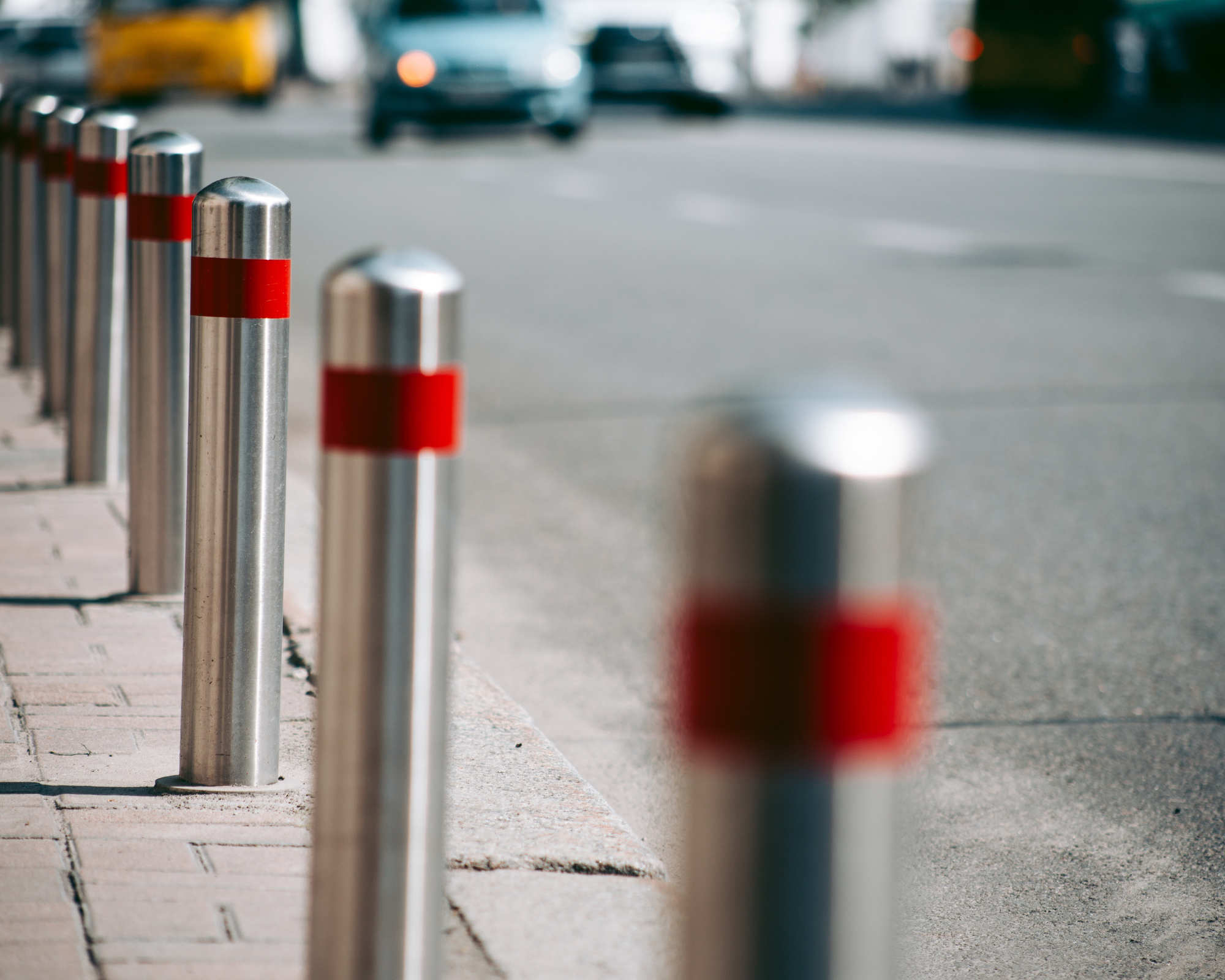 &nbsp;Row of cylindrical metal bollards with red bands, installed along a sidewalk.