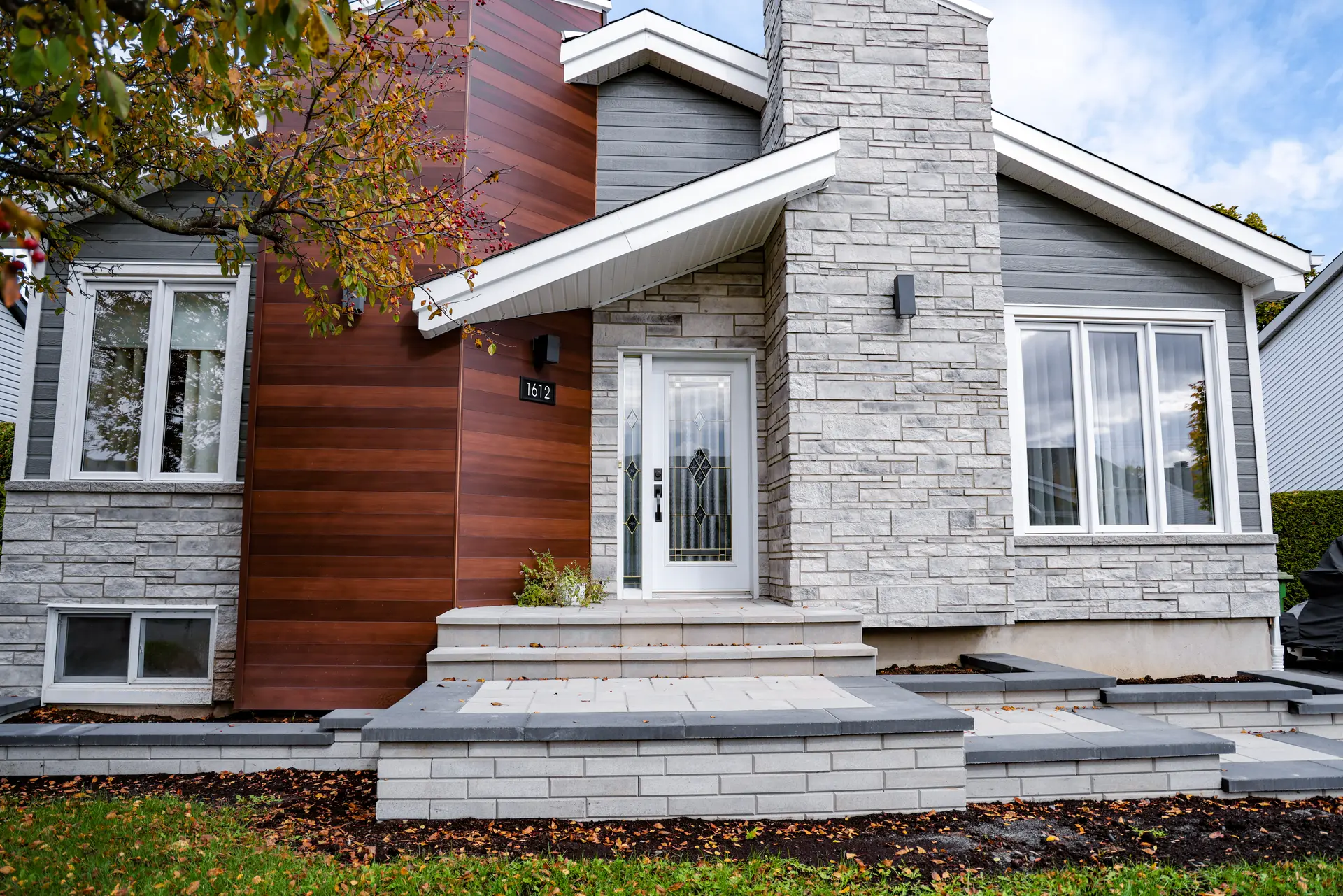 Renovated contemporary house façade with grey stone veneer, brown wood siding, white windows, a glazed front door, and masonry entrance steps