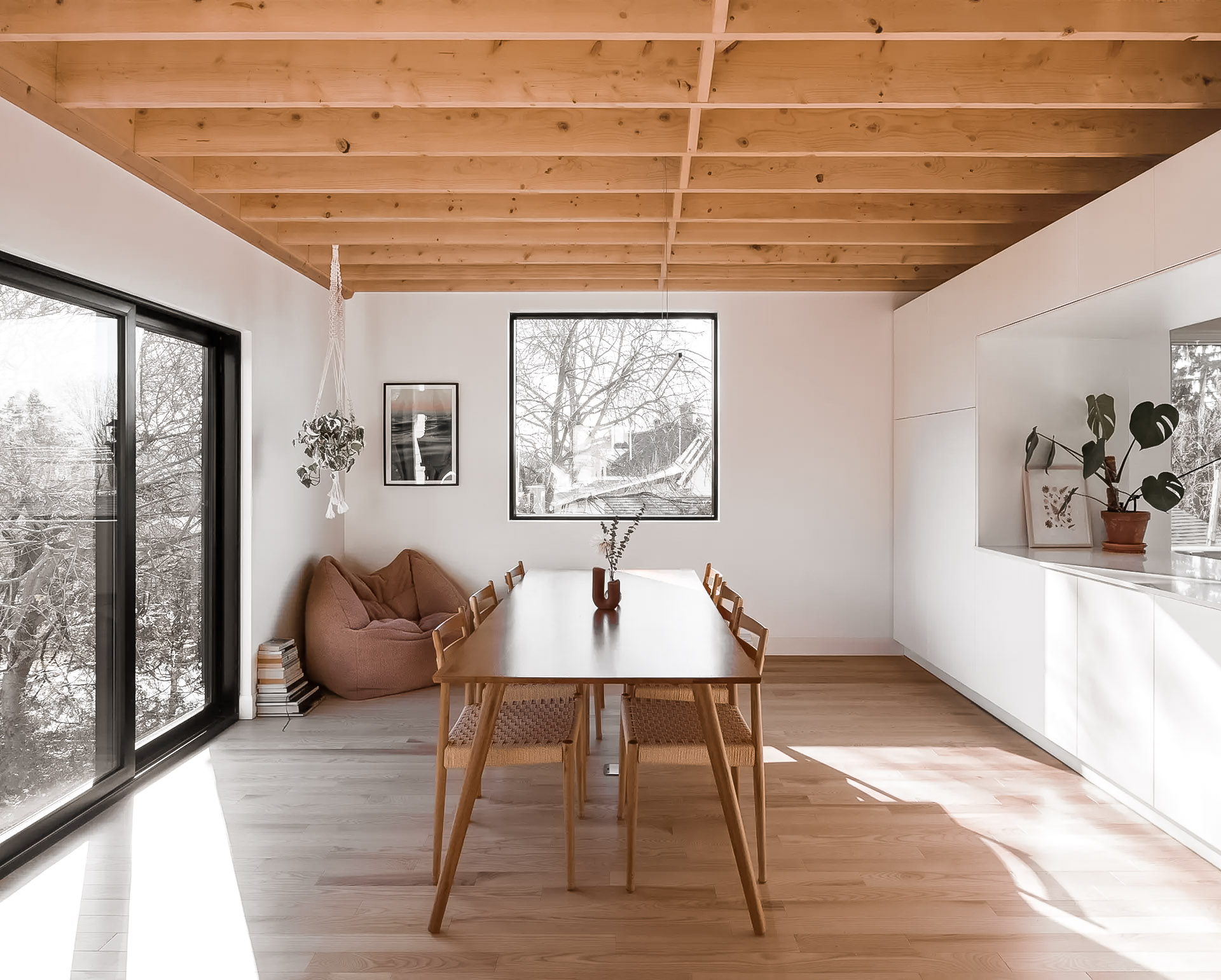 Salle à manger lumineuse avec plafond en bois apparent, grandes fenêtres noires, table en bois et décor minimaliste.