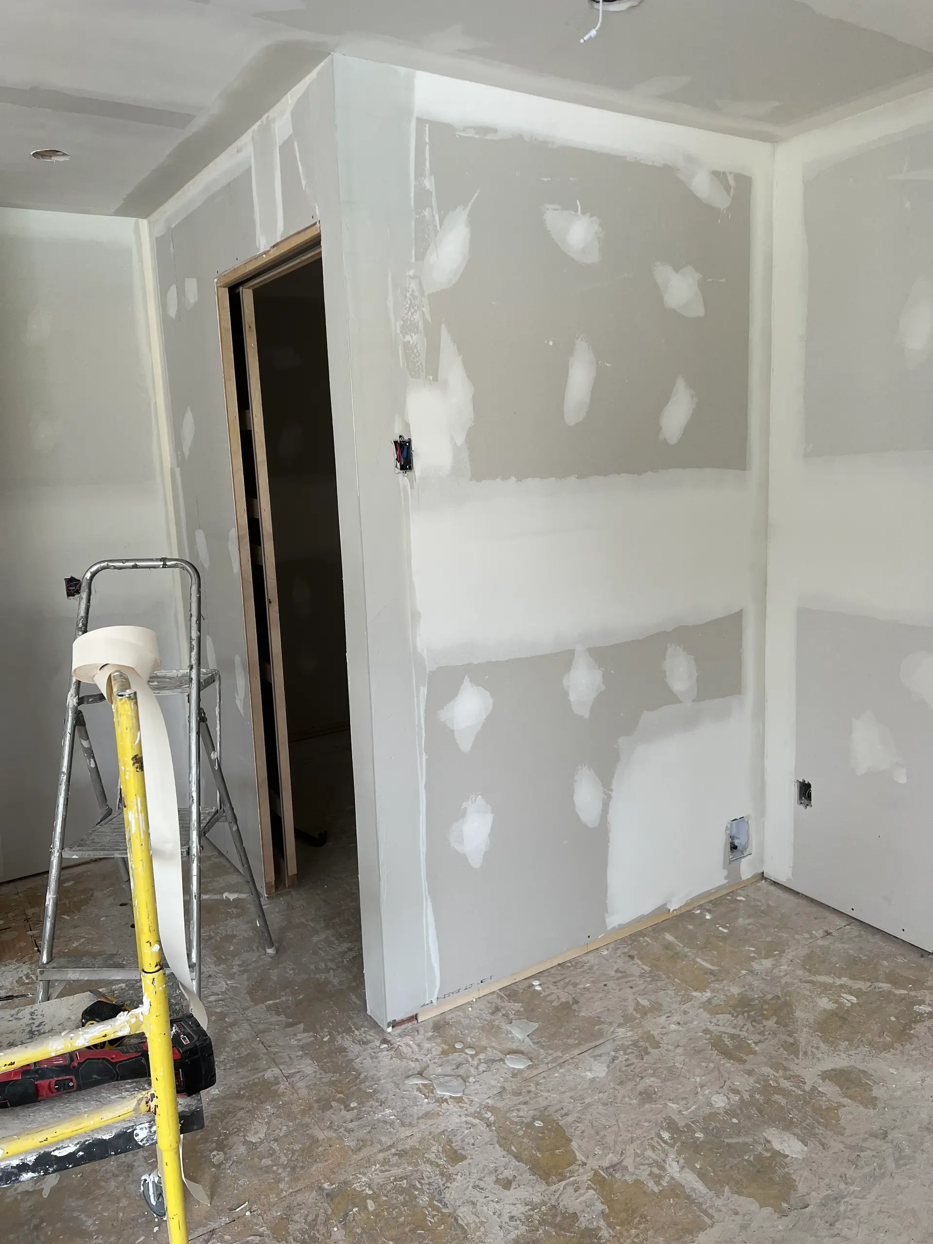 Room under renovation with taped drywall walls, electrical openings, a ladder, and plastering tools on the floor.