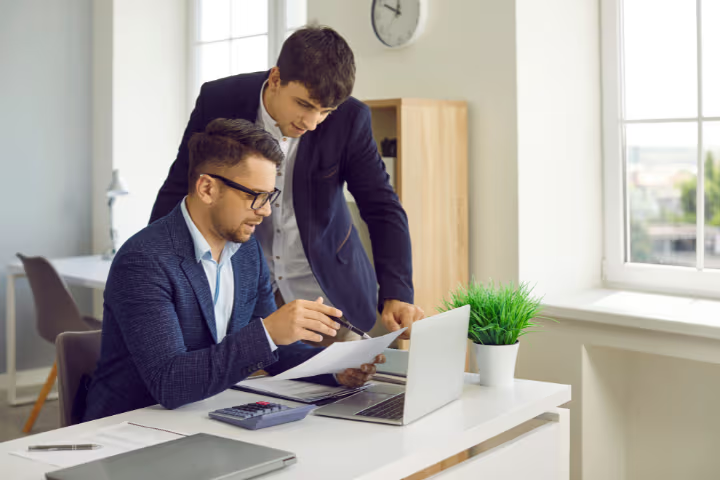 Two business professionals in suits reviewing financial documents with a laptop in a bright modern office.