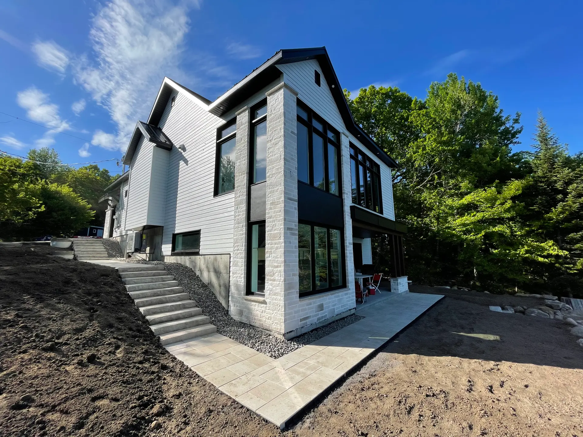 Maison contemporaine en bordure de forêt avec façade en briques pâles, grandes fenêtres noires et escalier sur terrain en pente