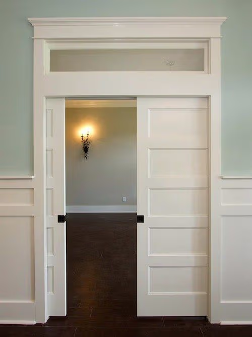 Portes coulissantes blanches encastrées avec moulures classiques, imposte vitrée et boiseries murales dans un intérieur élégant.
