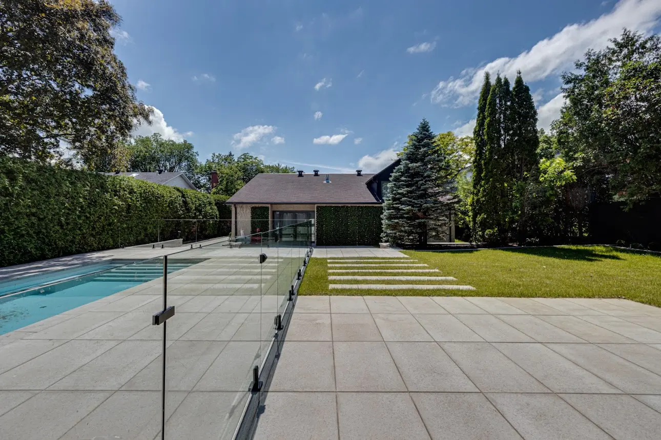 Contemporary backyard with inground pool, glass fence, slab terrace, landscaped lawn, mature hedges, trees and residential home