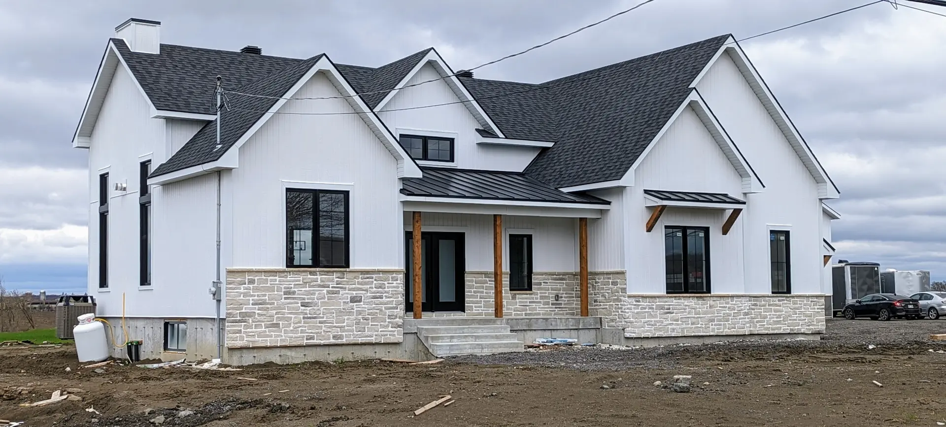 Maison neuve en construction avec revêtement blanc, base en pierre et toiture en bardeaux noirs sur un terrain non aménagé.
