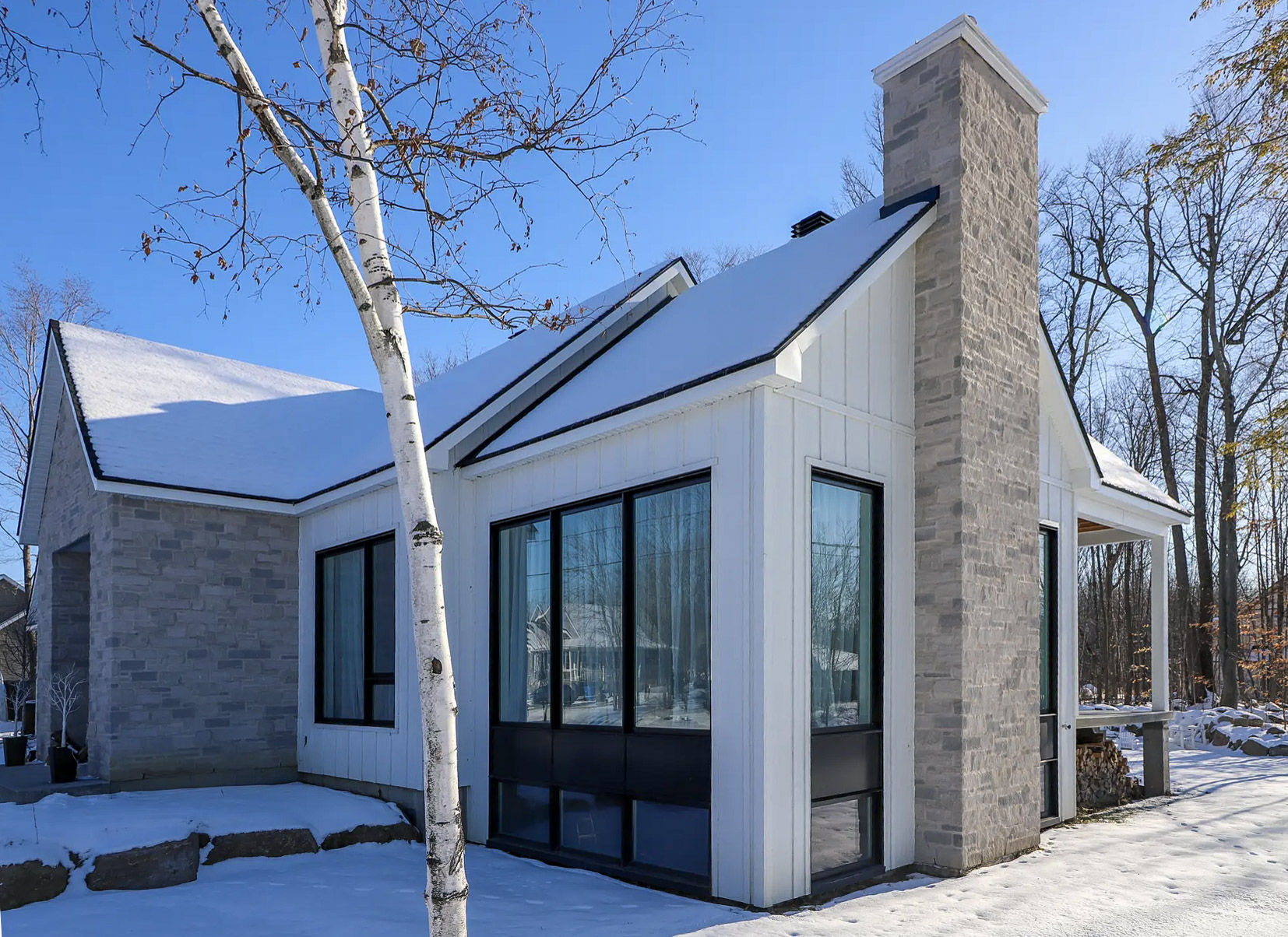 Modern home in winter with white wood and stone siding, large windows, and a stone chimney in a wooded environment.