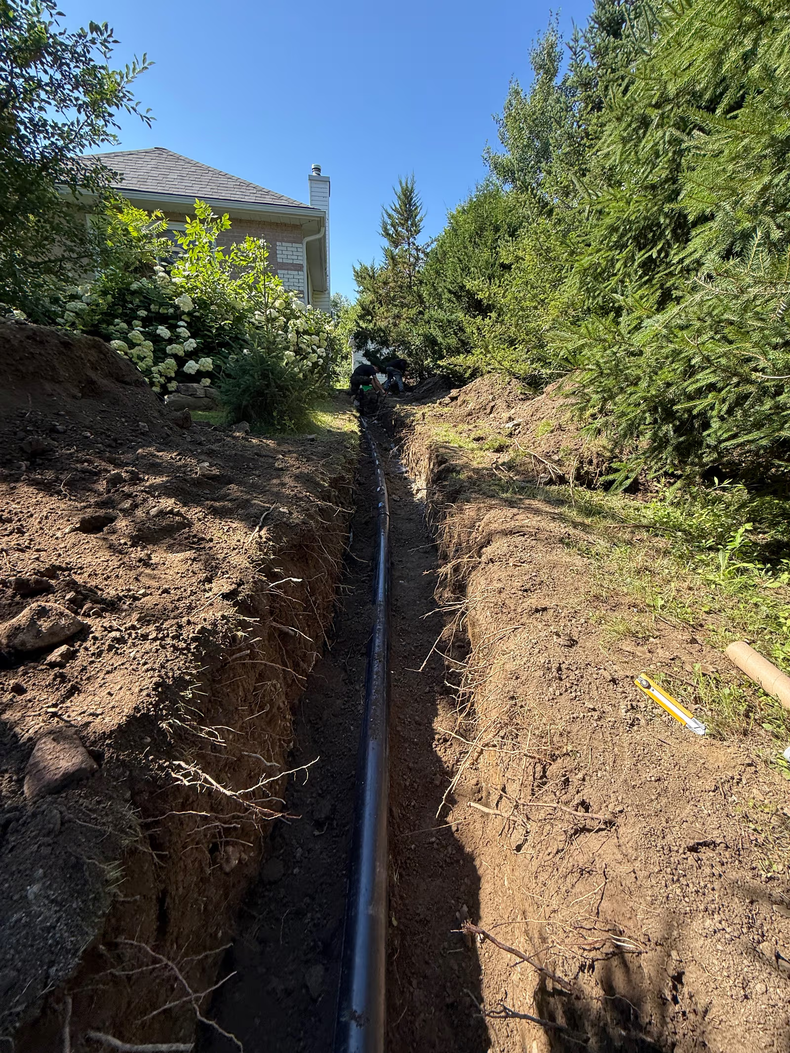 Tranchée d’excavation extérieure avec tuyau de drainage noir installé le long d’une maison résidentielle entourée d’arbres et d’aménagement paysager