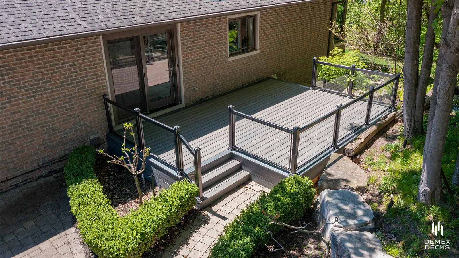 Composite deck with black aluminum and glass railing, accessed via a sliding door, surrounded by greenery and landscape pavers.