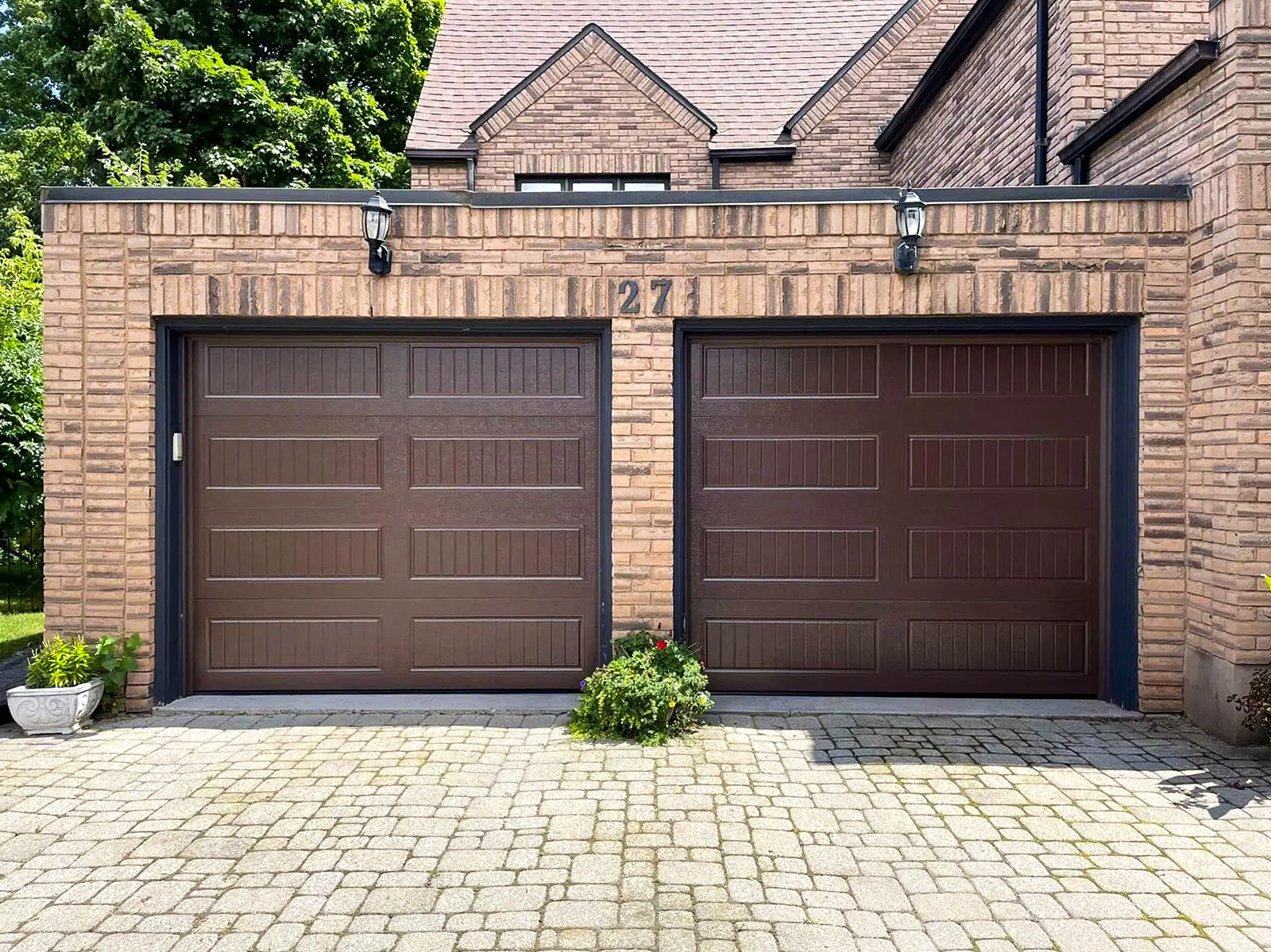 Garage résidentiel en brique avec deux portes brunes, luminaires extérieurs, pavé uni et aménagement paysager devant la façade.