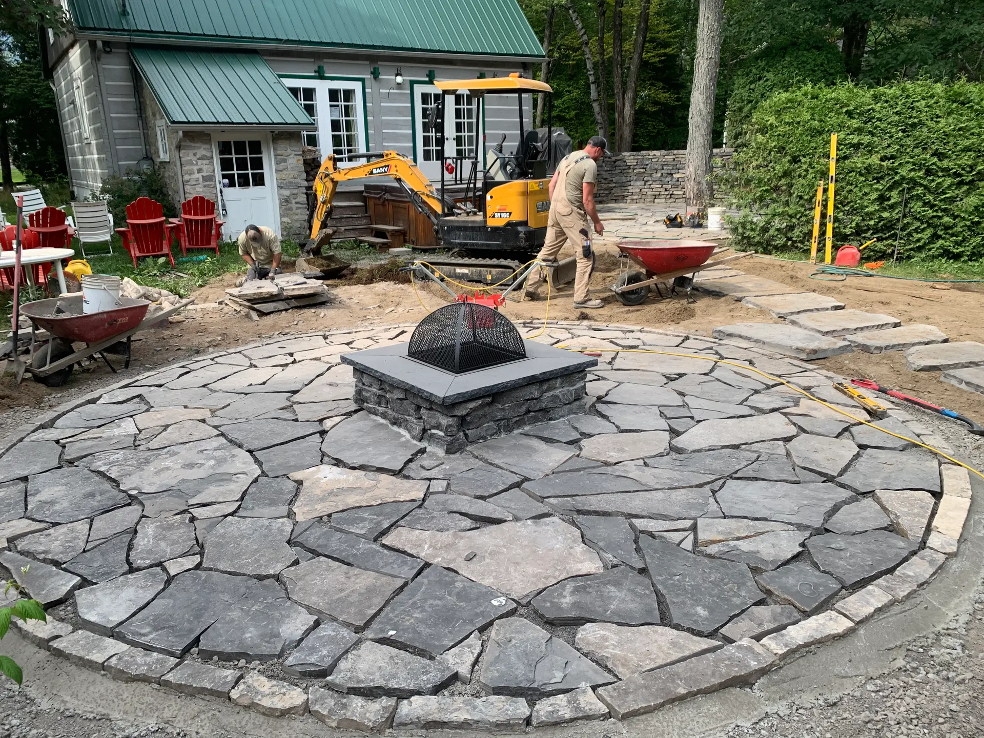 Installation of an outdoor fire pit on a natural stone surface in a landscaping project under construction in front of a wooden and green metal house.