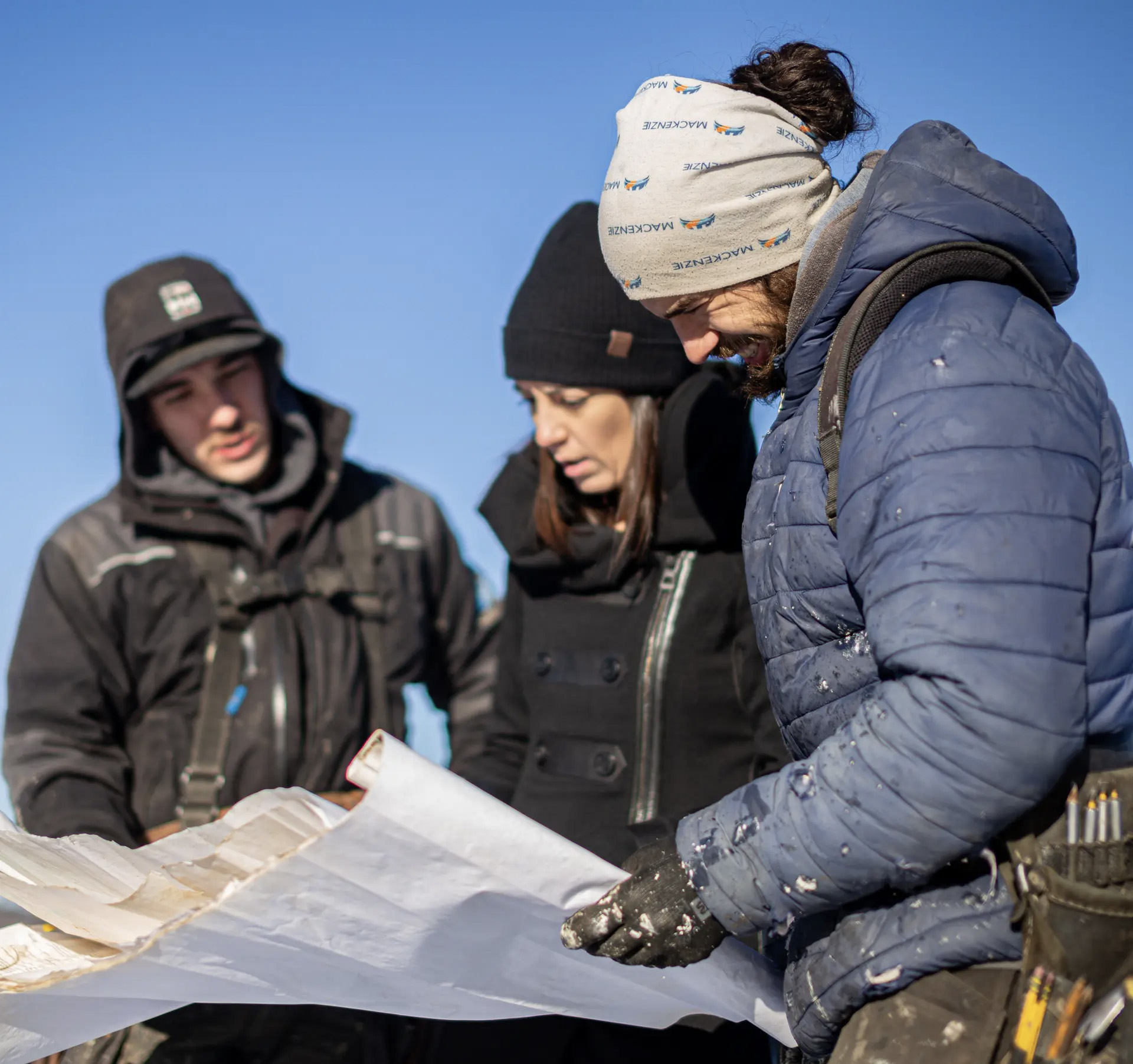 Trois personnes vêtues de vêtements de travail d’hiver examinent des plans de construction à l’extérieur par une journée ensoleillée, sous un ciel bleu dégagé.