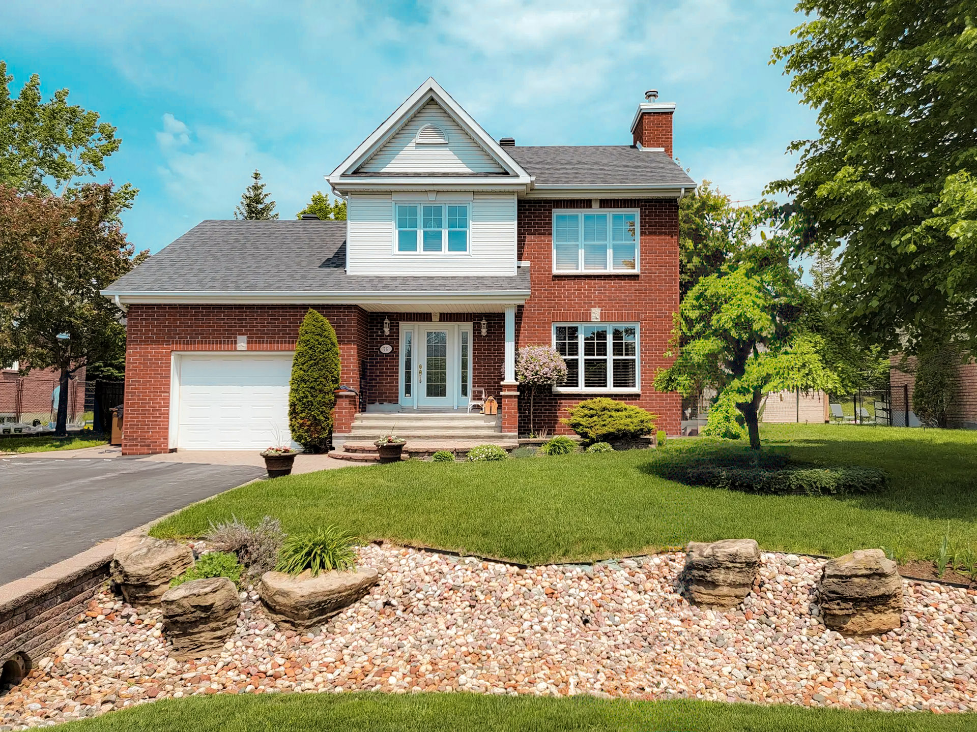 Red brick family home with integrated garage, shingled roof, manicured landscaping and decorative stone flowerbed