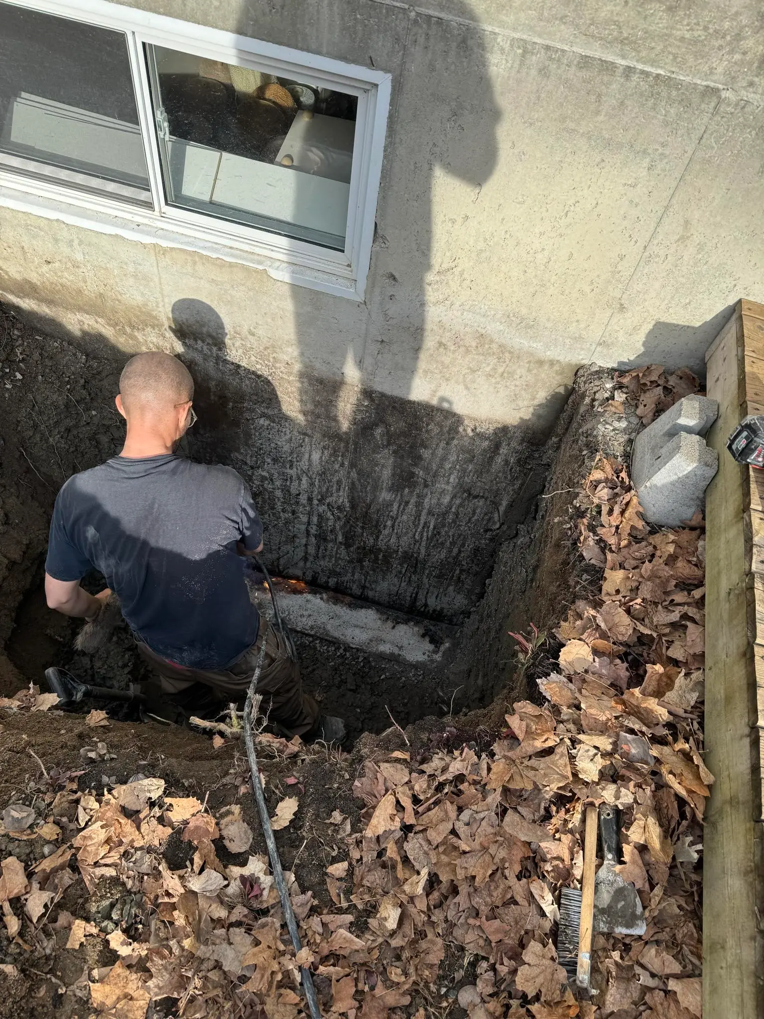 Worker in a foundation excavation near a basement window with exposed concrete wall, cleared soil and preparation for drainage and waterproofing