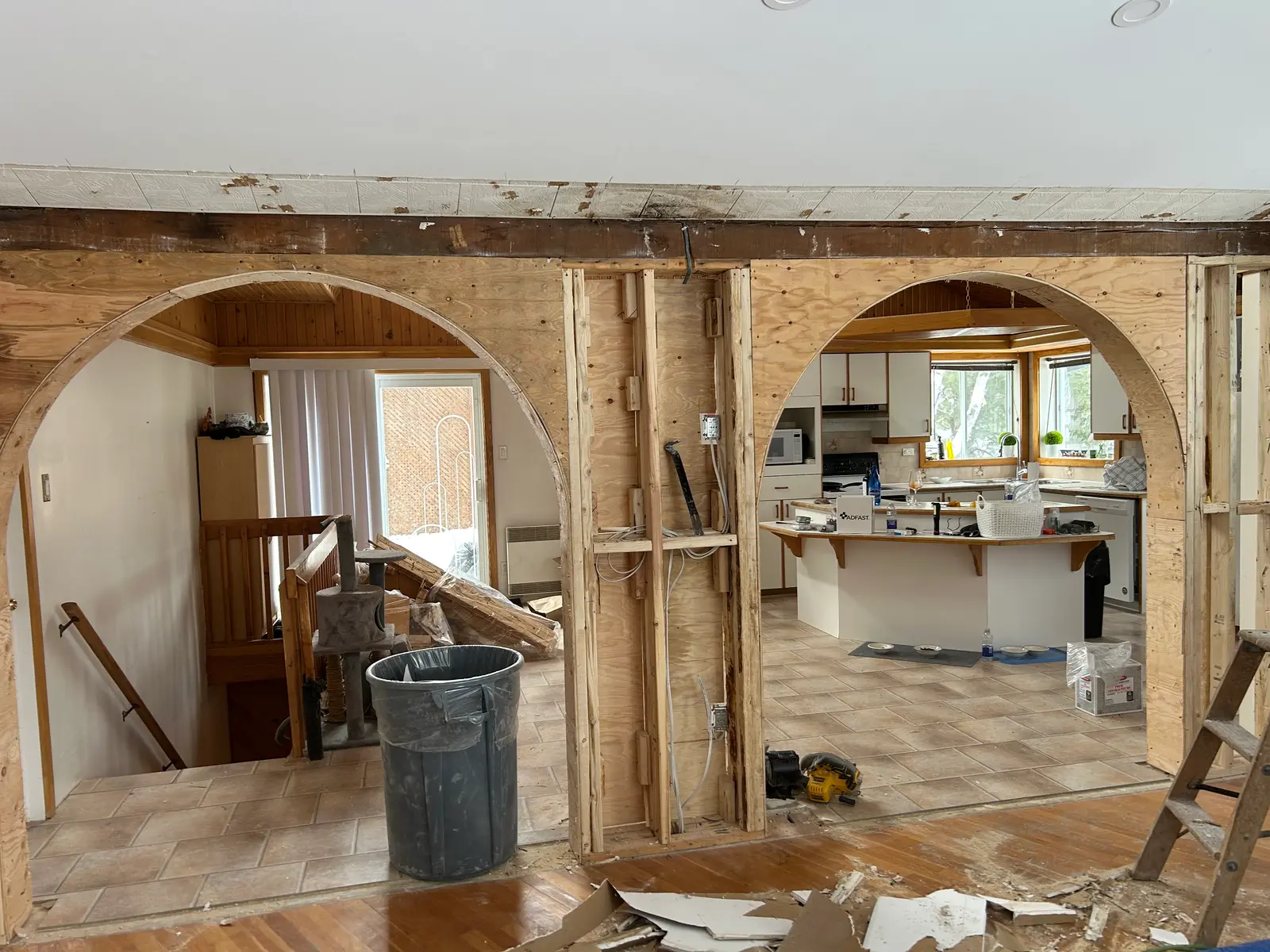 Intérieur de maison en rénovation avec mur partiellement démoli, grandes ouvertures cintrées, ossature en bois apparente et cuisine visible à l’arrière-plan.