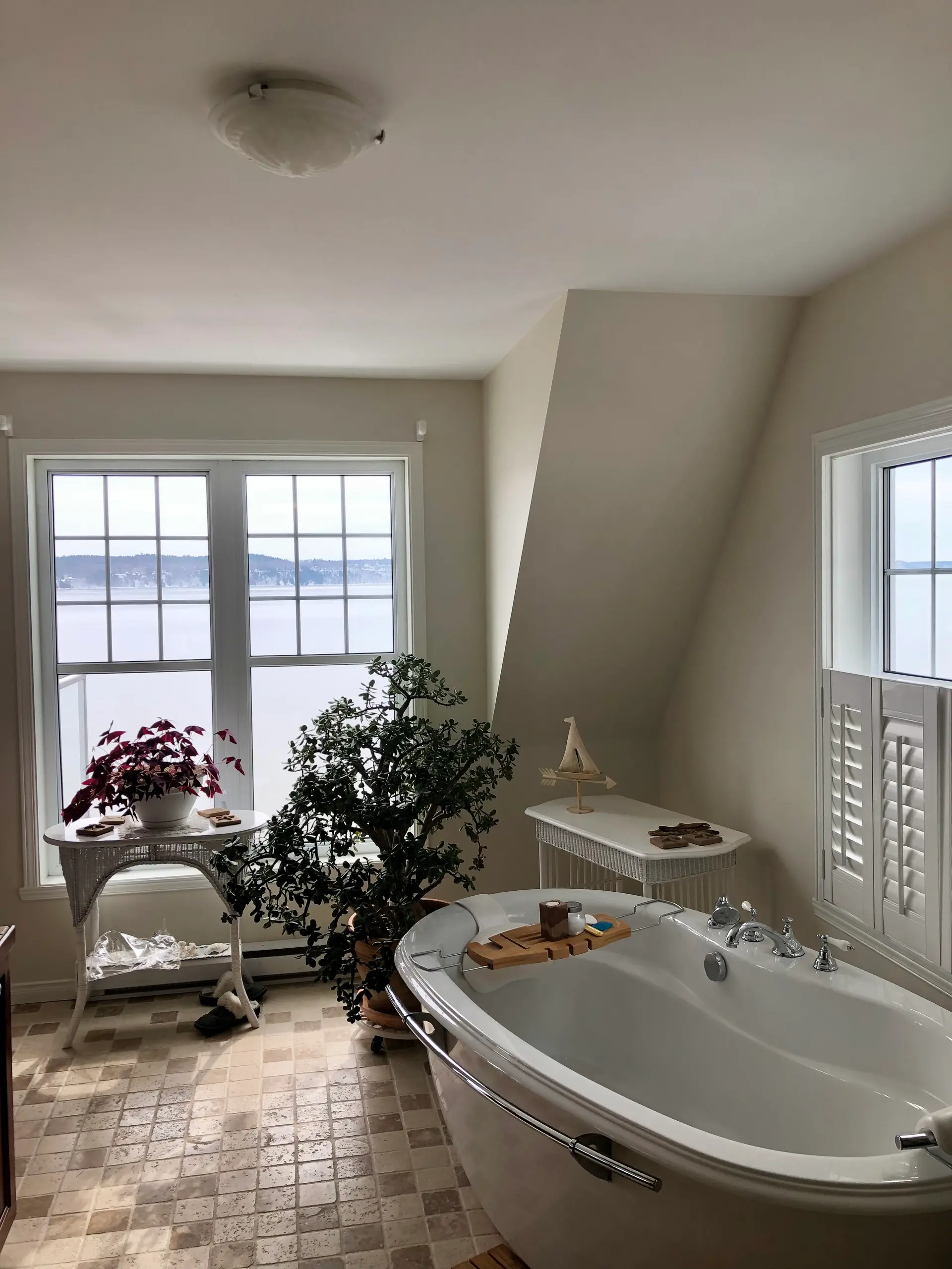 Salle de bain lumineuse avec baignoire autoportante, carrelage en pierre naturelle et grandes fenêtres avec vue sur un paysage extérieur. Mobilier en rotin blanc et décoration végétale apaisante.