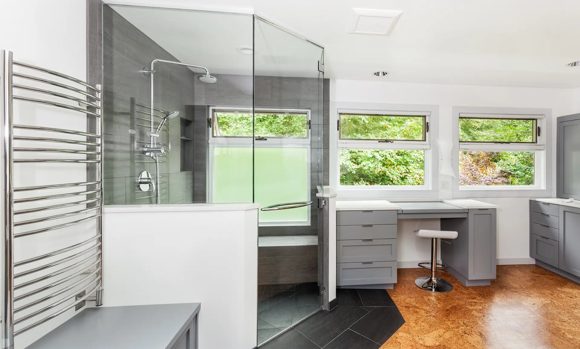 Salle de bain moderne avec douche en verre, plancher en liège, mobilier gris et grandes fenêtres lumineuses