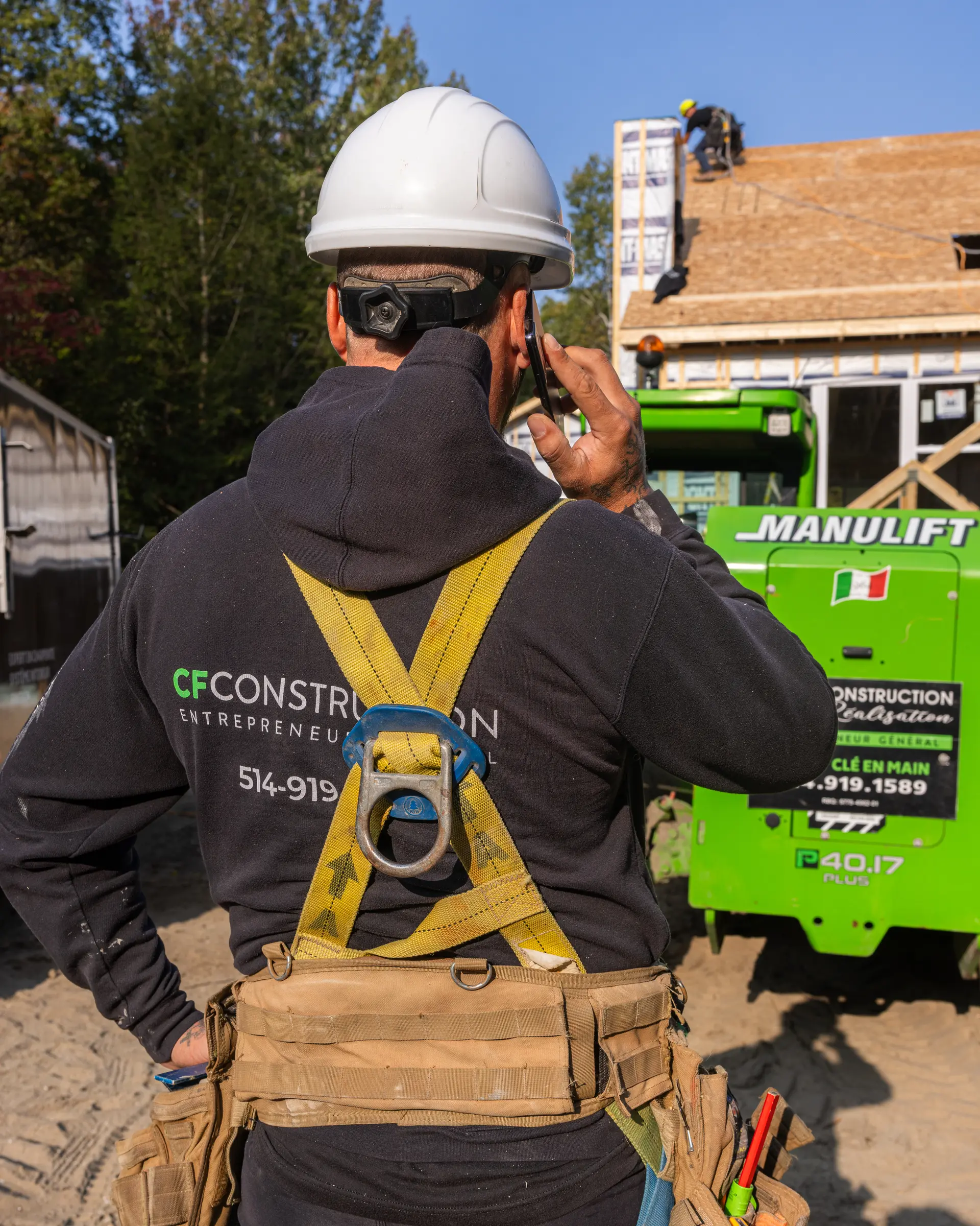 Travailleur de la construction portant un harnais de sécurité et un casque, en communication téléphonique sur un chantier avec des travaux de toiture en arrière-plan. /&nbsp;Construction worker wearing a safety harness and helmet, talking on the phone at a job site with roofing work in the background.