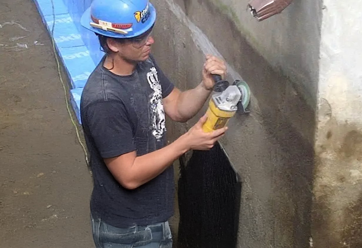 Worker wearing a blue hard hat using a grinder on a concrete foundation wall during waterproofing work
