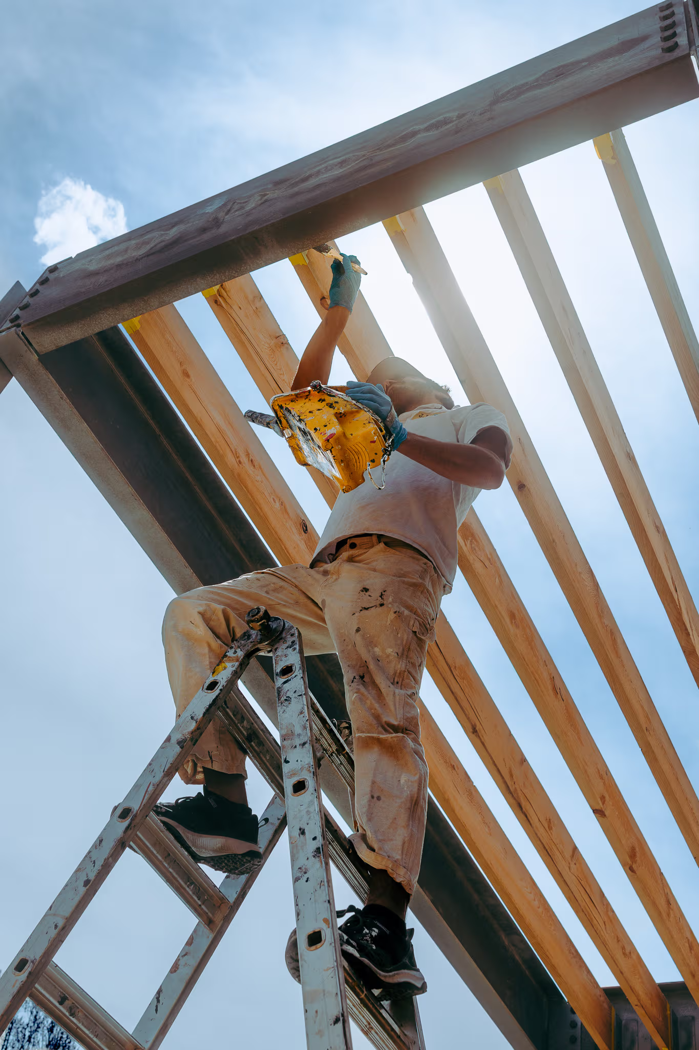 Peintre en bâtiment sur une échelle appliquant de la peinture sur une structure en bois extérieure sous un ciel bleu /&nbsp;House painter on a ladder applying paint to an outdoor wooden structure under a blue sky
