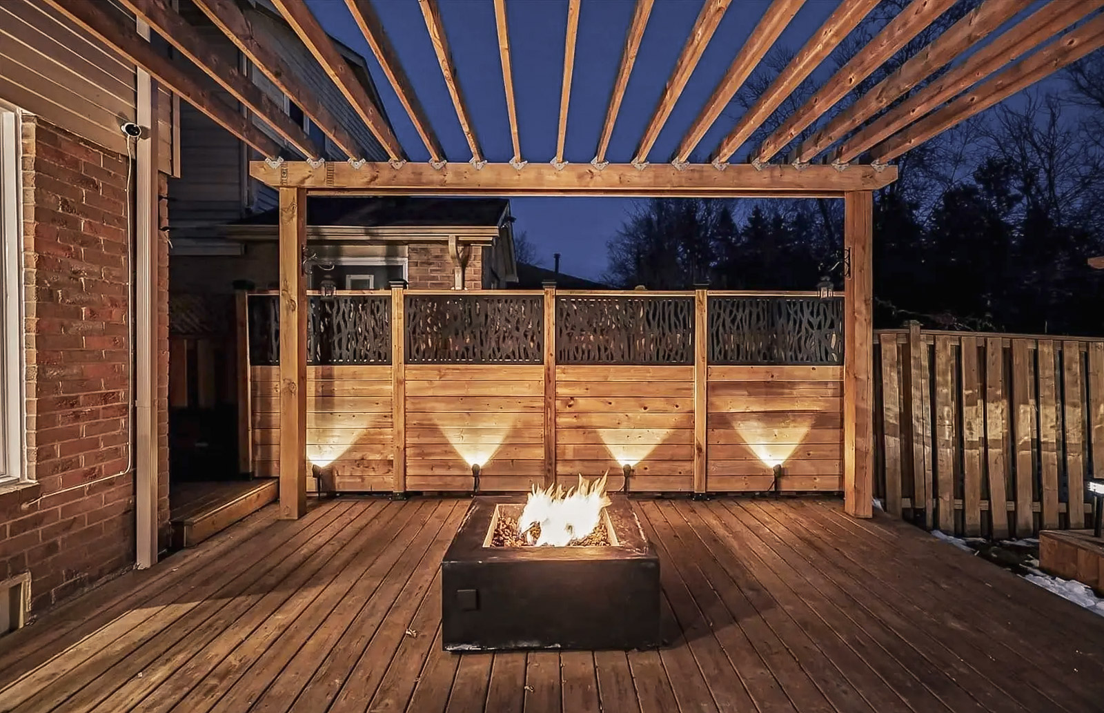 Terrasse en bois avec pergola et foyer extérieur illuminé, entourée d’une clôture en bois décorative, idéale pour des soirées chaleureuses en plein air.