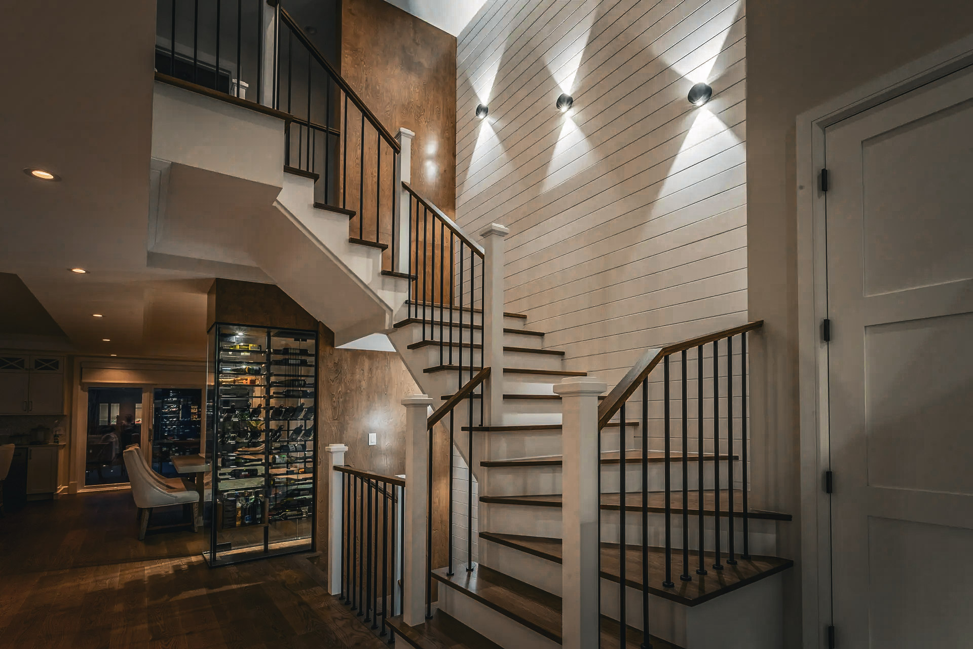 Intérieur moderne avec escalier en bois et métal noir, éclairage mural, panneaux de bois, cave à vin encastrée, et sous-sol aménagé.