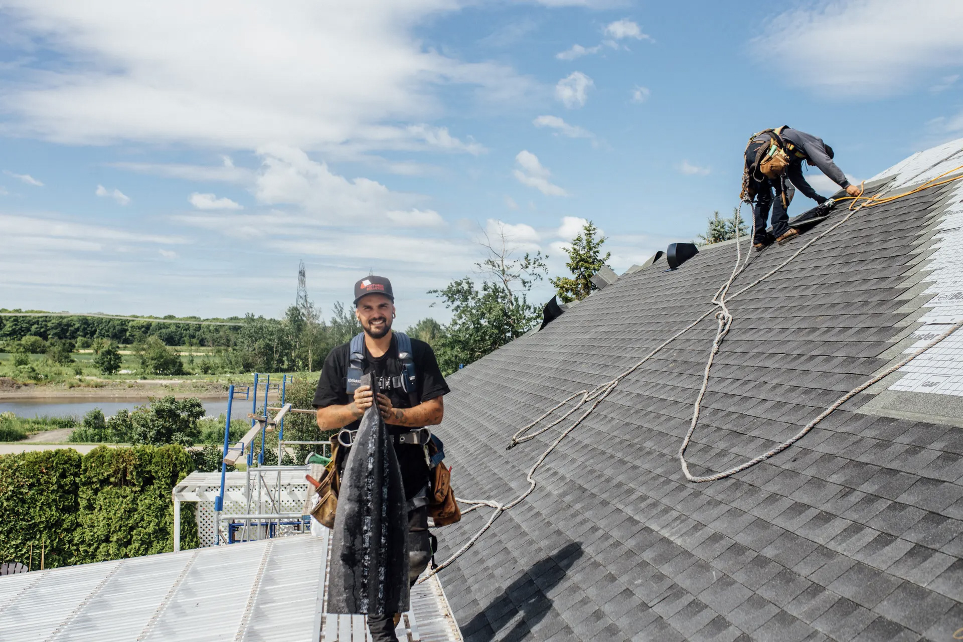 Deux couvreurs travaillent sur un toit en bardeaux sous un ciel bleu l’un sourit à la caméra en tenant une sortie de toit, tandis que l’autre fixe les bardeaux à l’aide de cordes de sécurité.