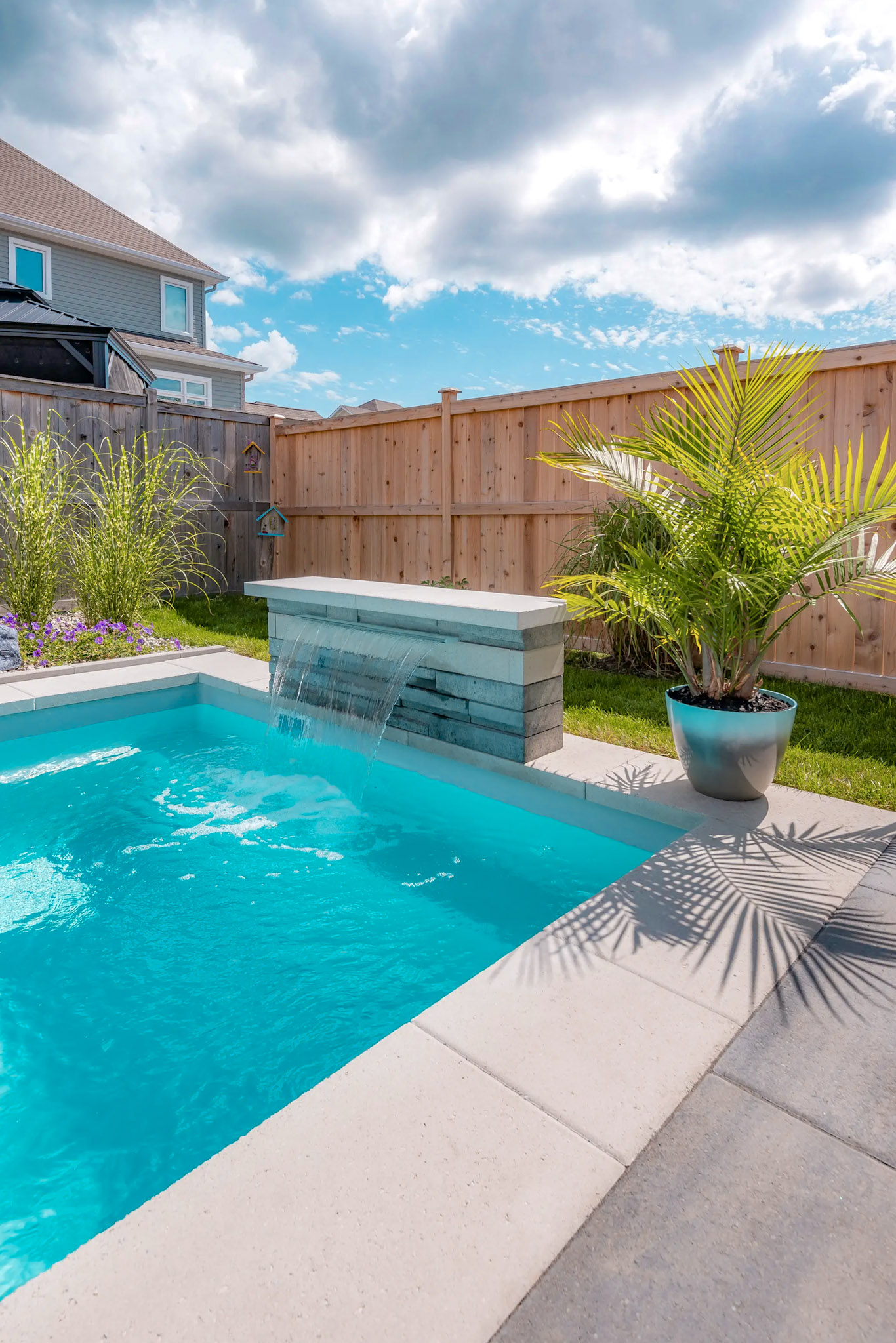 Inground pool with decorative stone waterfall and composite wood fence in a landscaped residential yard