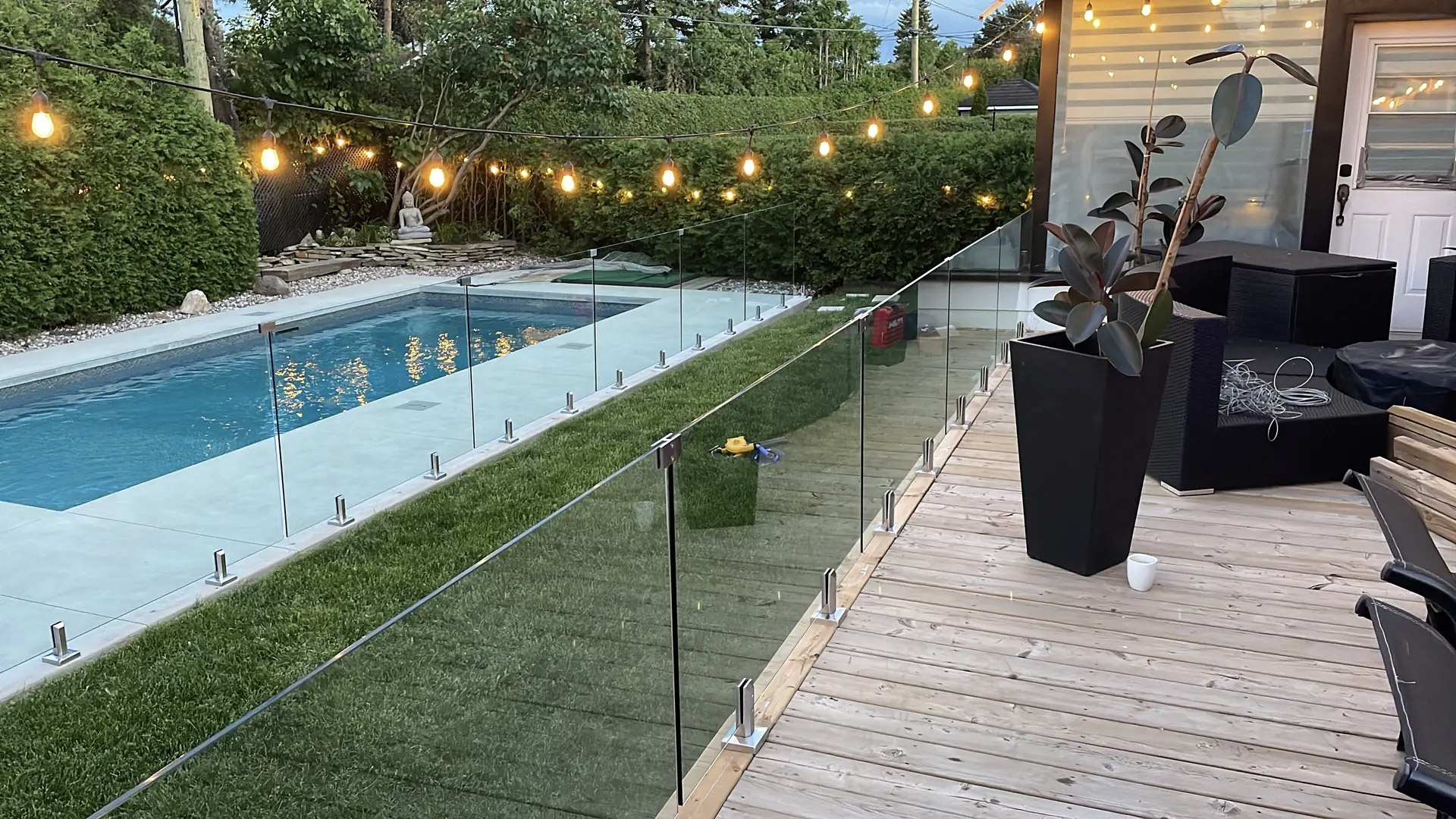 Backyard with in-ground pool, glass fence, wooden deck, outdoor lighting and mature hedges.