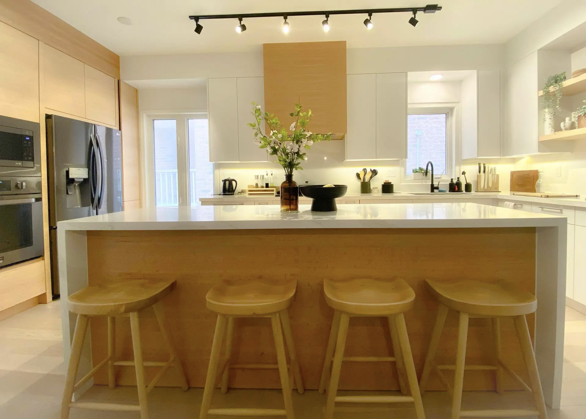 Bright modern kitchen with a large central island, white countertop, light wood cabinetry, four stools, and track lighting.