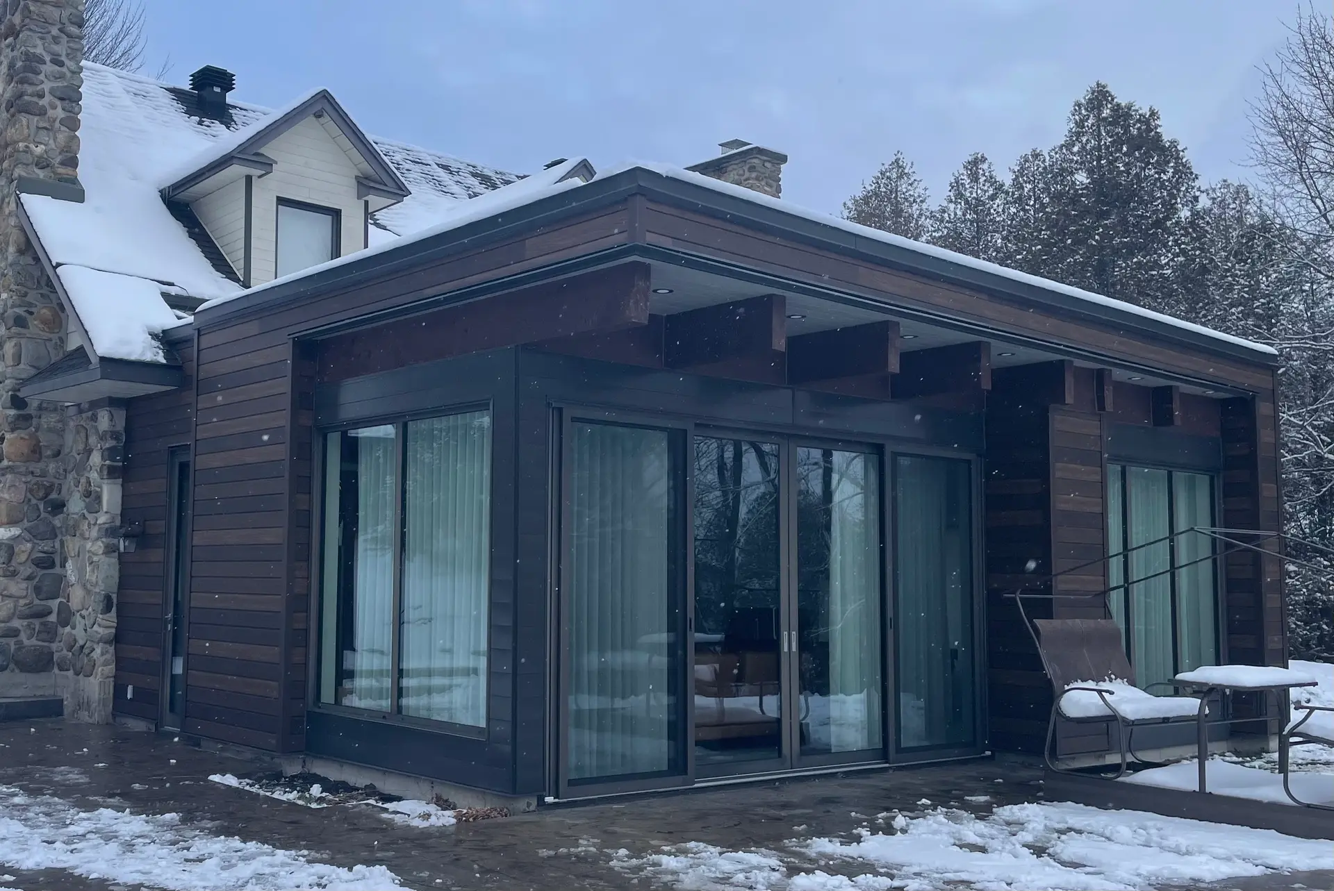 Modern house extension with dark wood siding, large sliding glass doors, flat roof, and snow-covered patio.